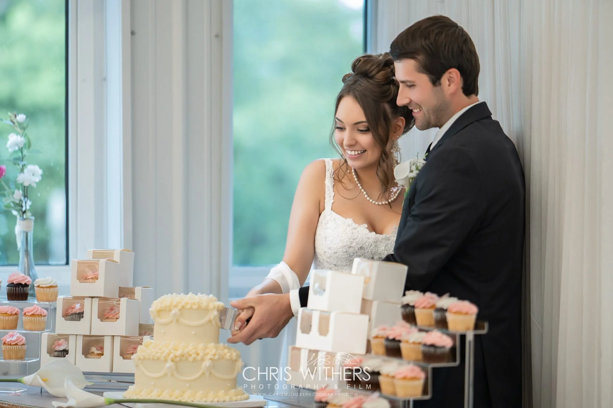 A bride and groom at a wedding reception cutting a wedding cake, with desserts and cupcakes on display.