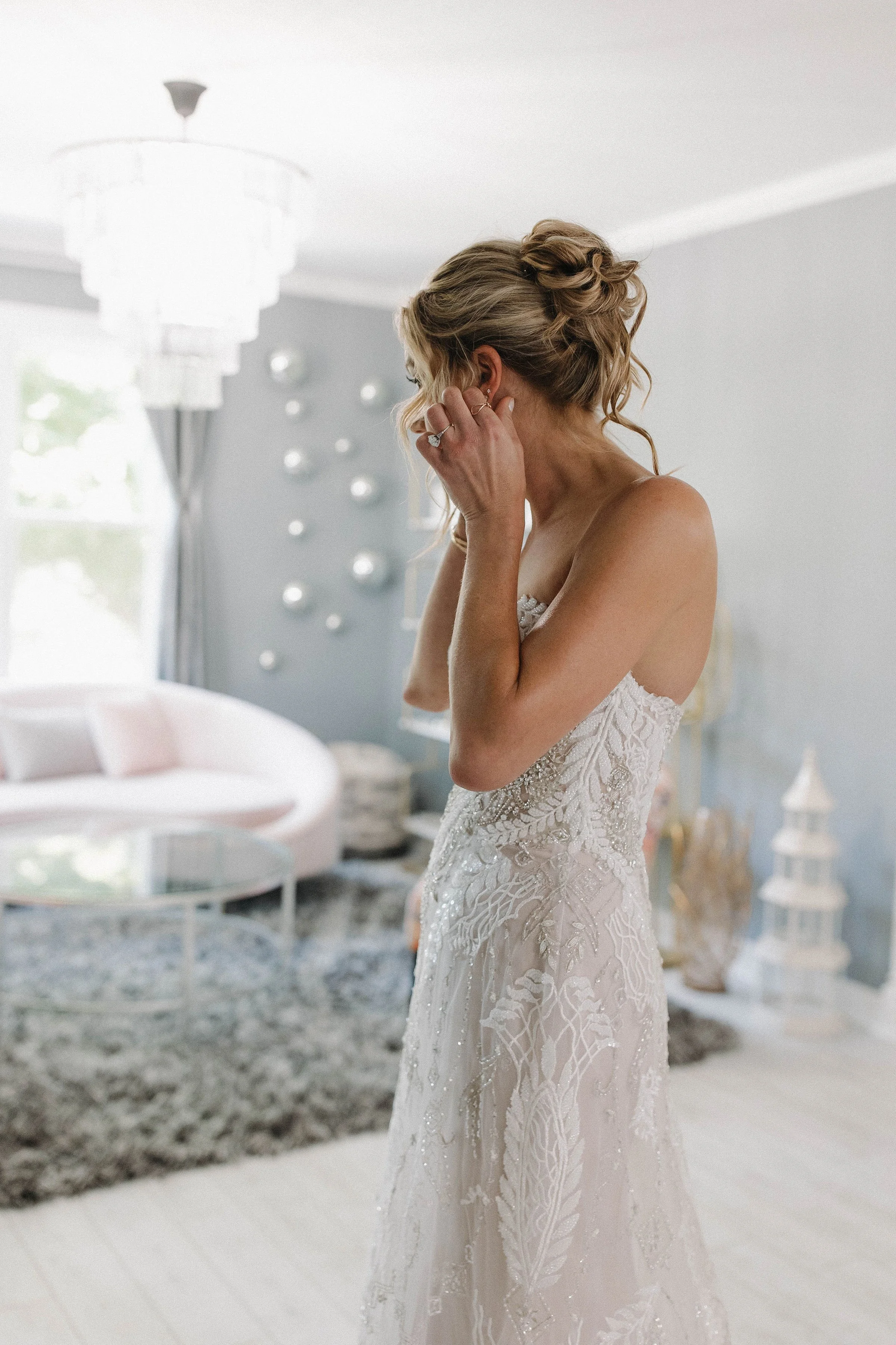 A woman in a wedding dress putting in an earring in a brightly lit room decorated with modern art pieces.