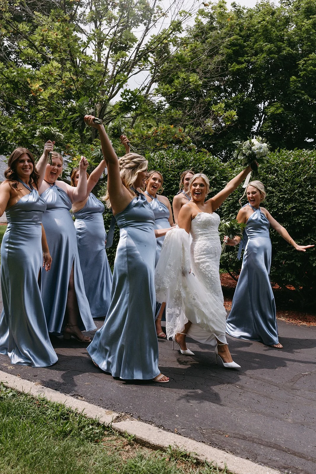 A bride in a white wedding gown joyfully walking with bridesmaids in matching blue satin dresses, celebrating outdoors with trees in the background.