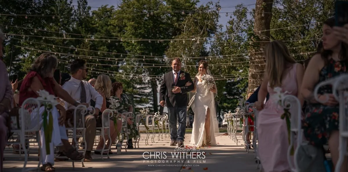 A bride walking down the aisle with her escort at an outdoor wedding ceremony, surrounded by seated guests under string lights and trees.