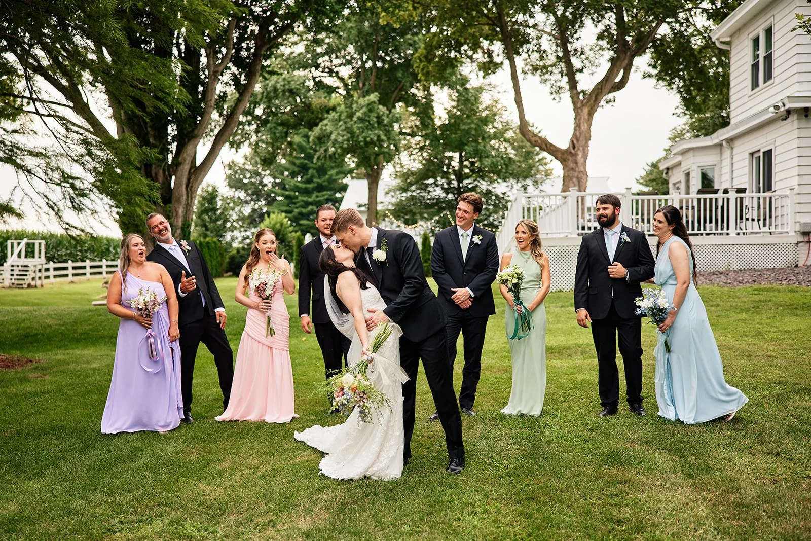 A wedding party outdoors with the bride and groom sharing a kiss, surrounded by bridesmaids and groomsmen, on a green lawn with trees and a white house in the background.