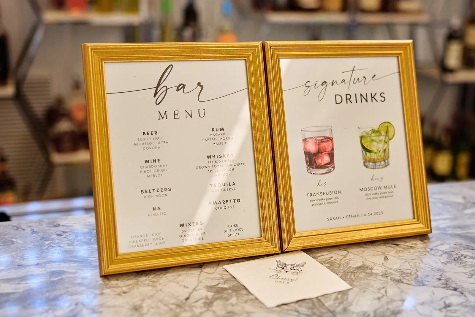 Two framed signage boards on a marble countertop display a bar menu and signature drinks at an event. One board lists drinks by category, and the other features images of two cocktails with their names, ingredients, and a wedding date of June 14, 202