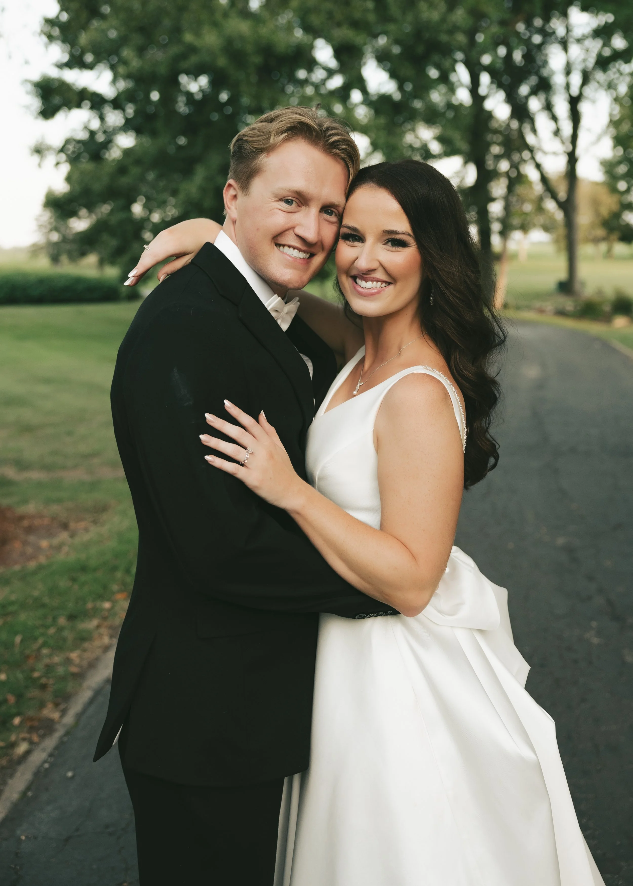 A newlywed couple is smiling and holding each other outdoors in a park with trees in the background, dressed in wedding attire.