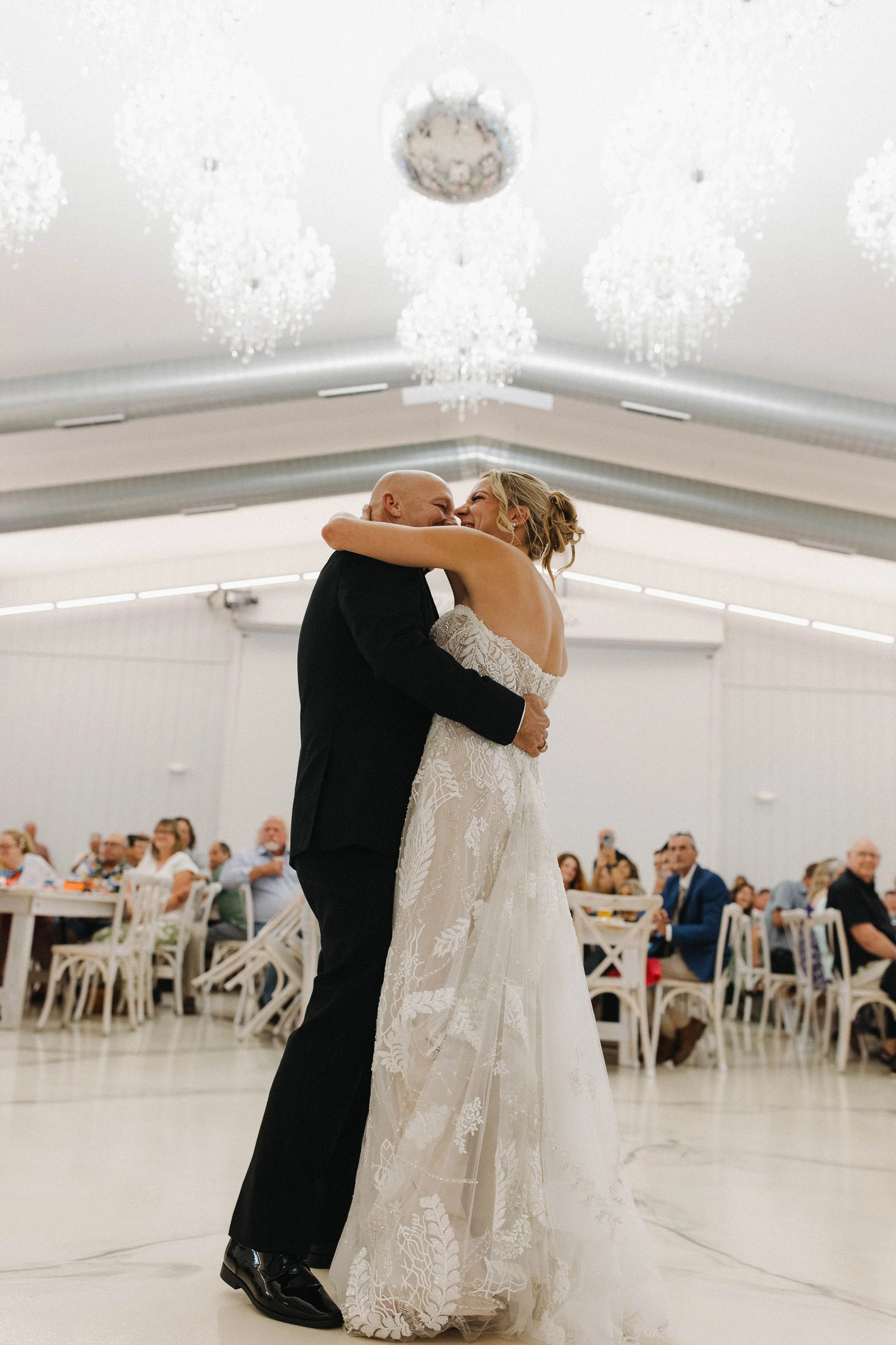 A bride and a groom sharing a dance at their wedding reception. The bride is in a white lace dress, and the groom is in a black suit. Guests are seated at tables in the background, and the ceiling has white chandeliers.