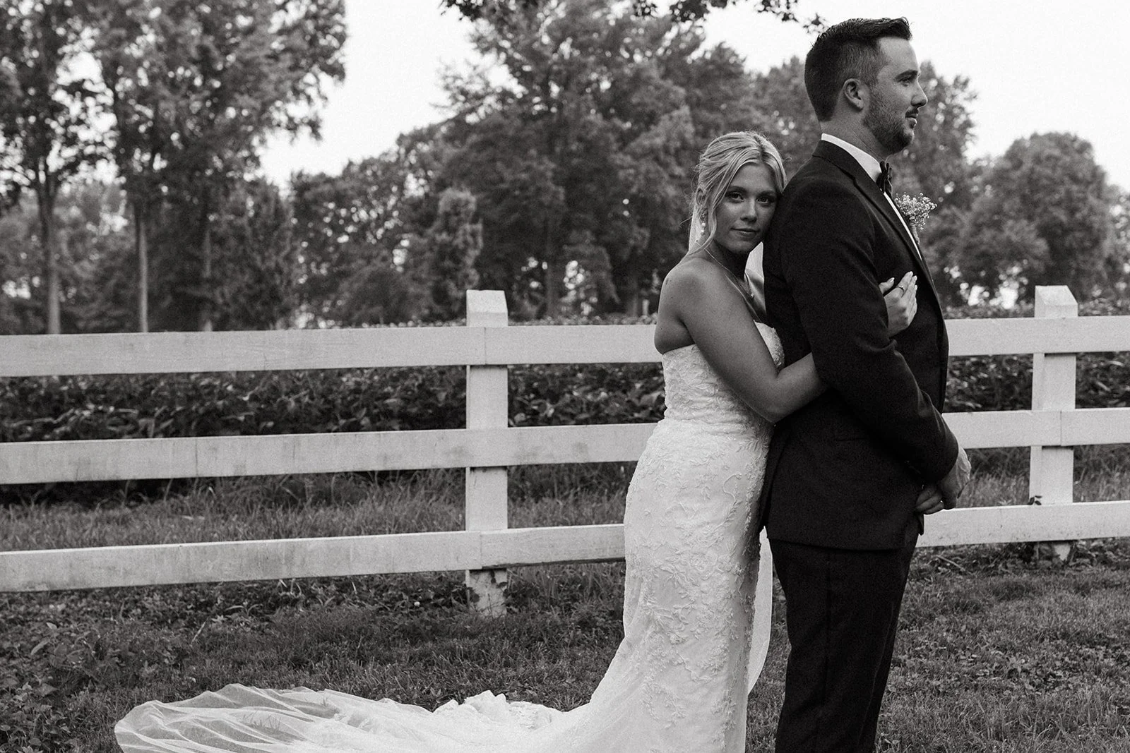 A black and white photograph of a bride and groom standing outdoors. The bride is hugging the groom from behind and wearing a strapless lace wedding gown with a train, while the groom is dressed in a black tuxedo. They are standing in front of a whit