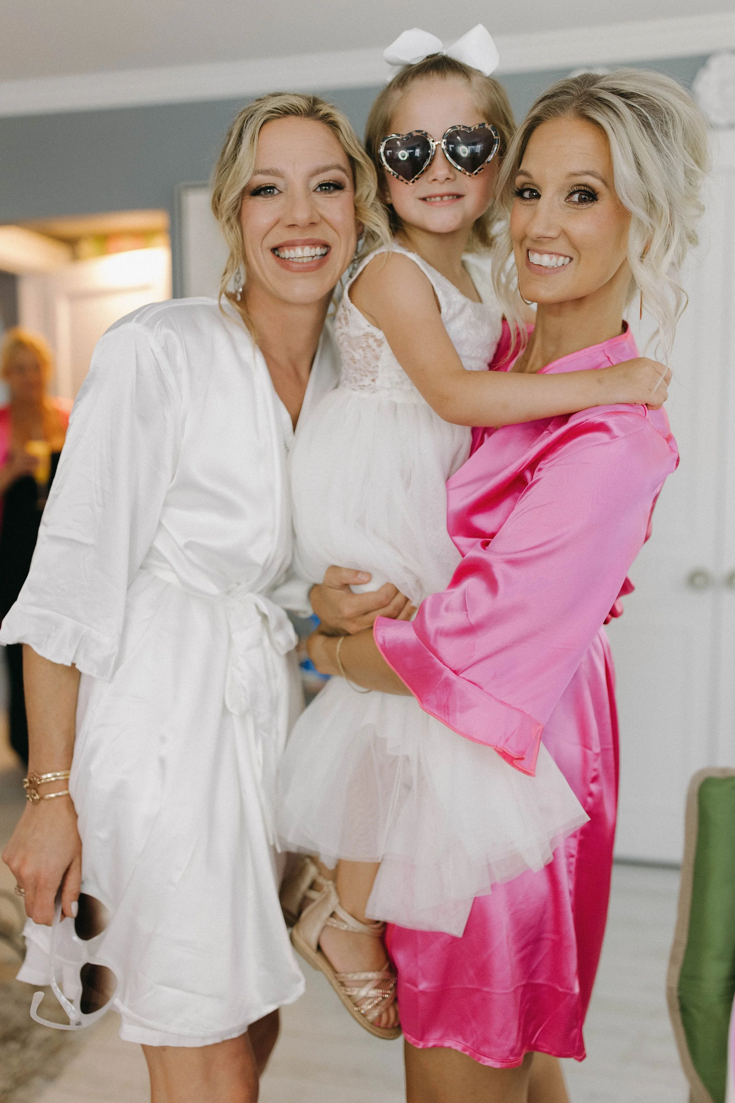 Three women, two adults and a child, smiling and posing together indoors. The child wears heart-shaped sunglasses and a white lace dress, held by a woman in a pink satin robe. Another woman in a white satin robe stands beside them.