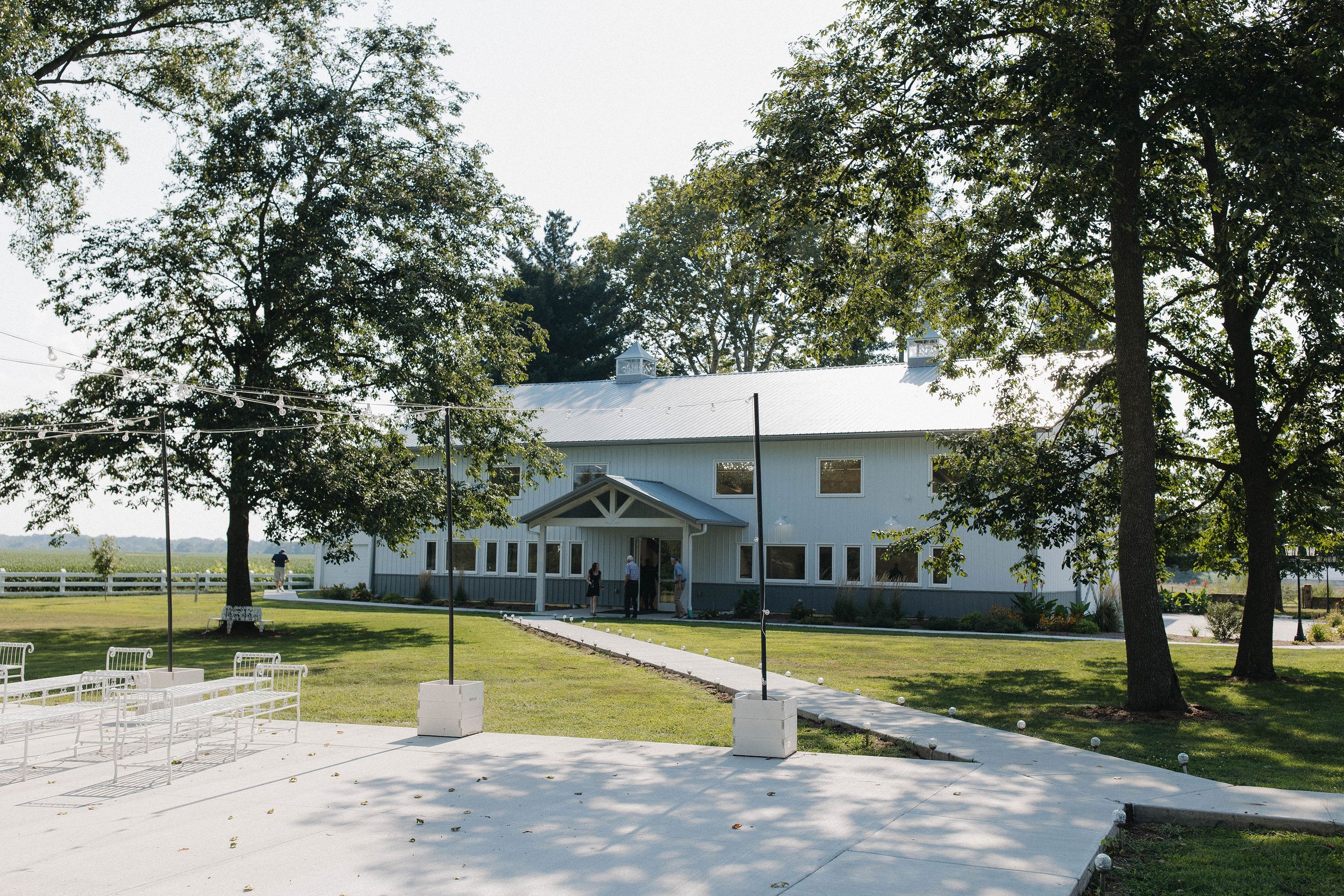 A white barn-style building with a metal roof surrounded by green trees and grass, with a concrete pathway leading to the entrance.
