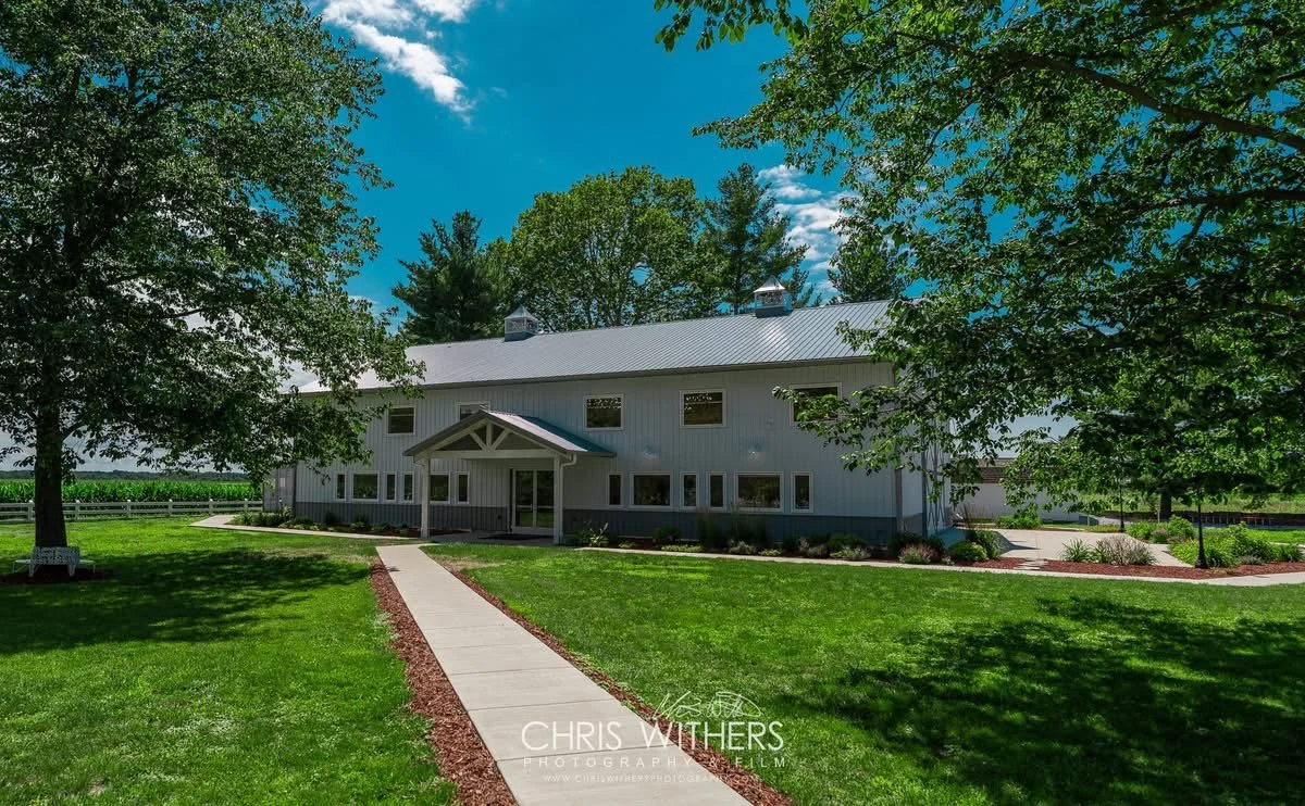 A large white barn with a metal roof surrounded by green grass and trees, with a clear blue sky overhead.