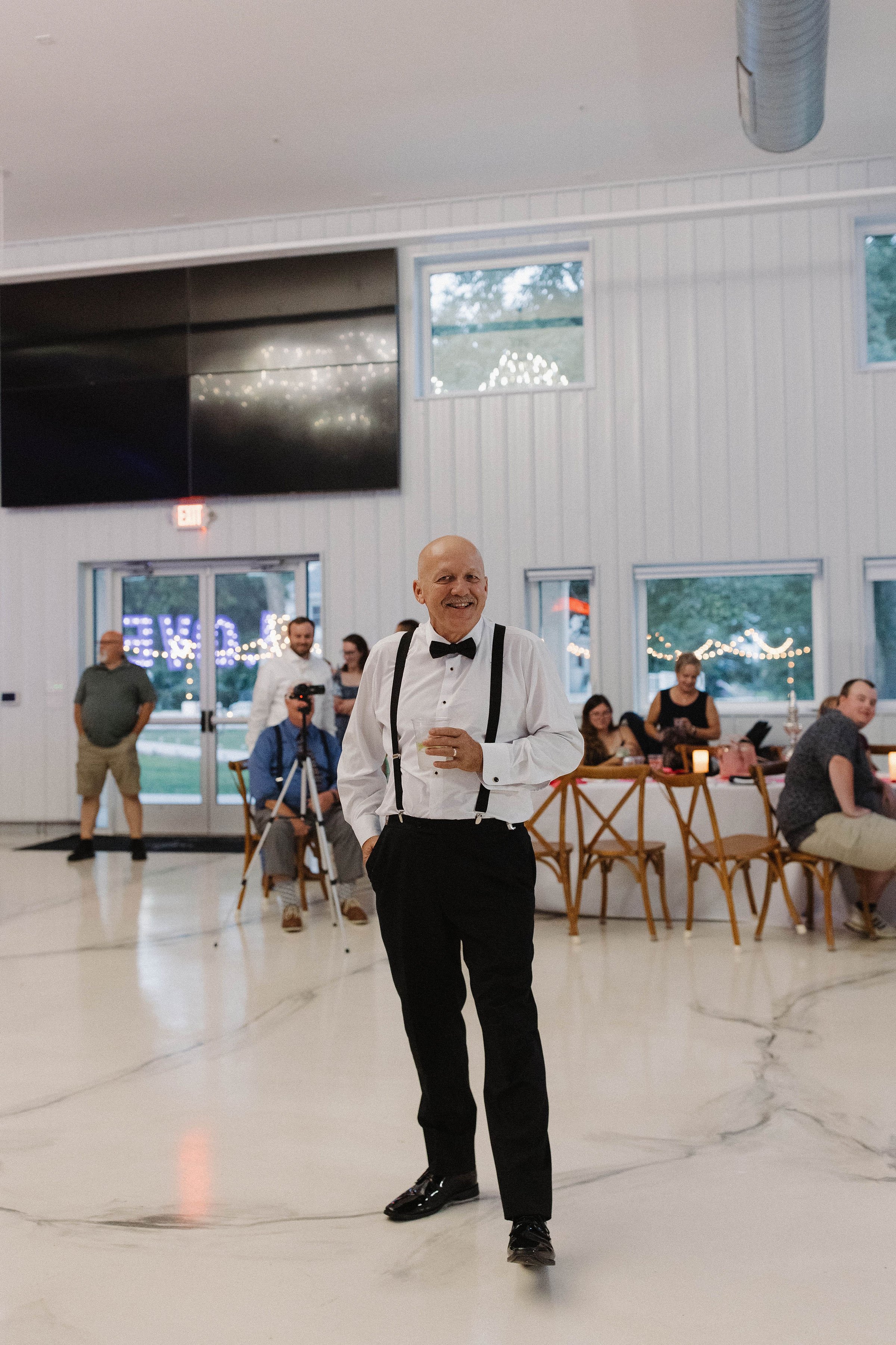 A man in a tuxedo with suspenders and a bow tie, smiling, standing in a reception hall with other guests and decorations.