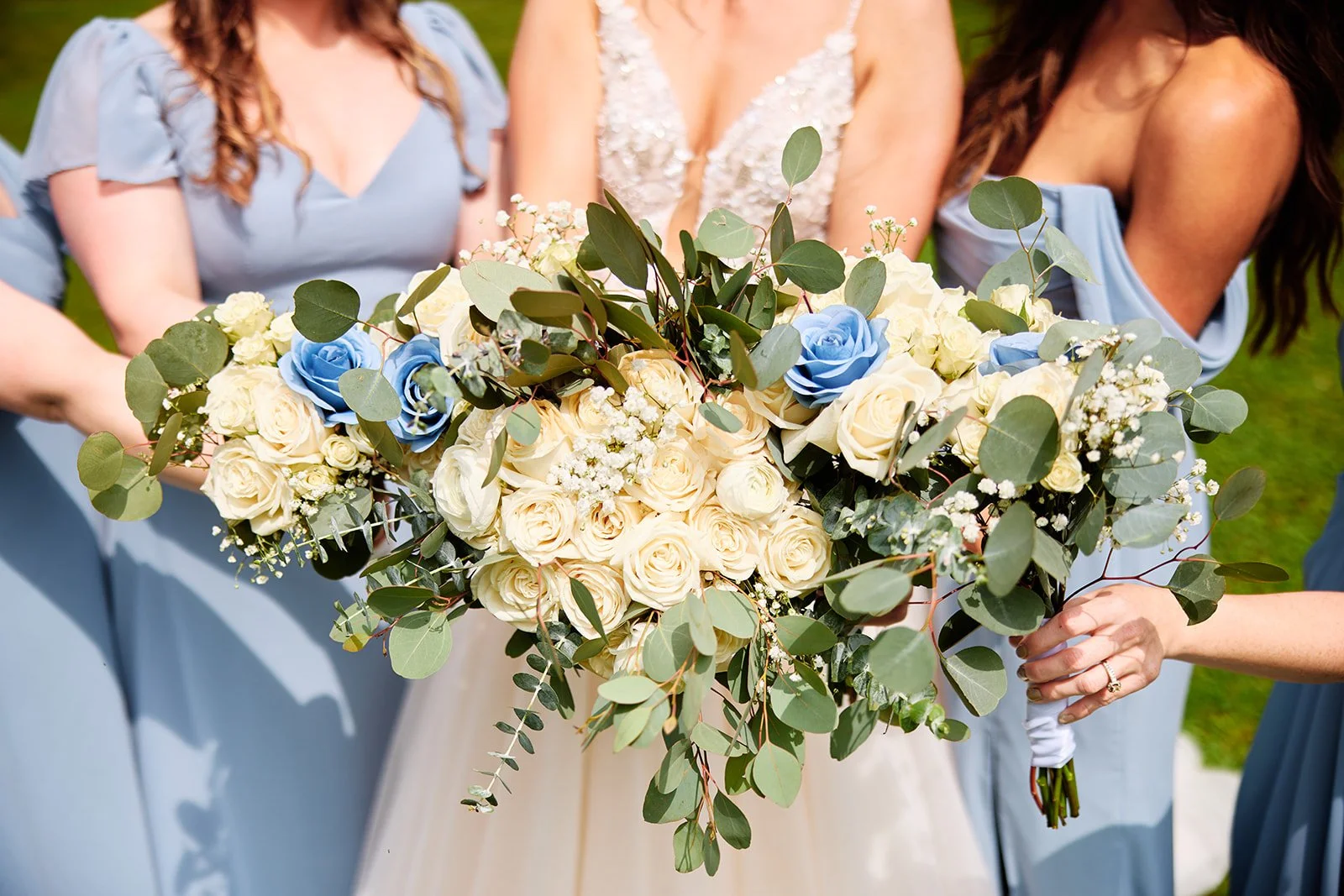 A bride holding a large bouquet of white and blue roses with greenery, surrounded by women in light blue dresses at a wedding.