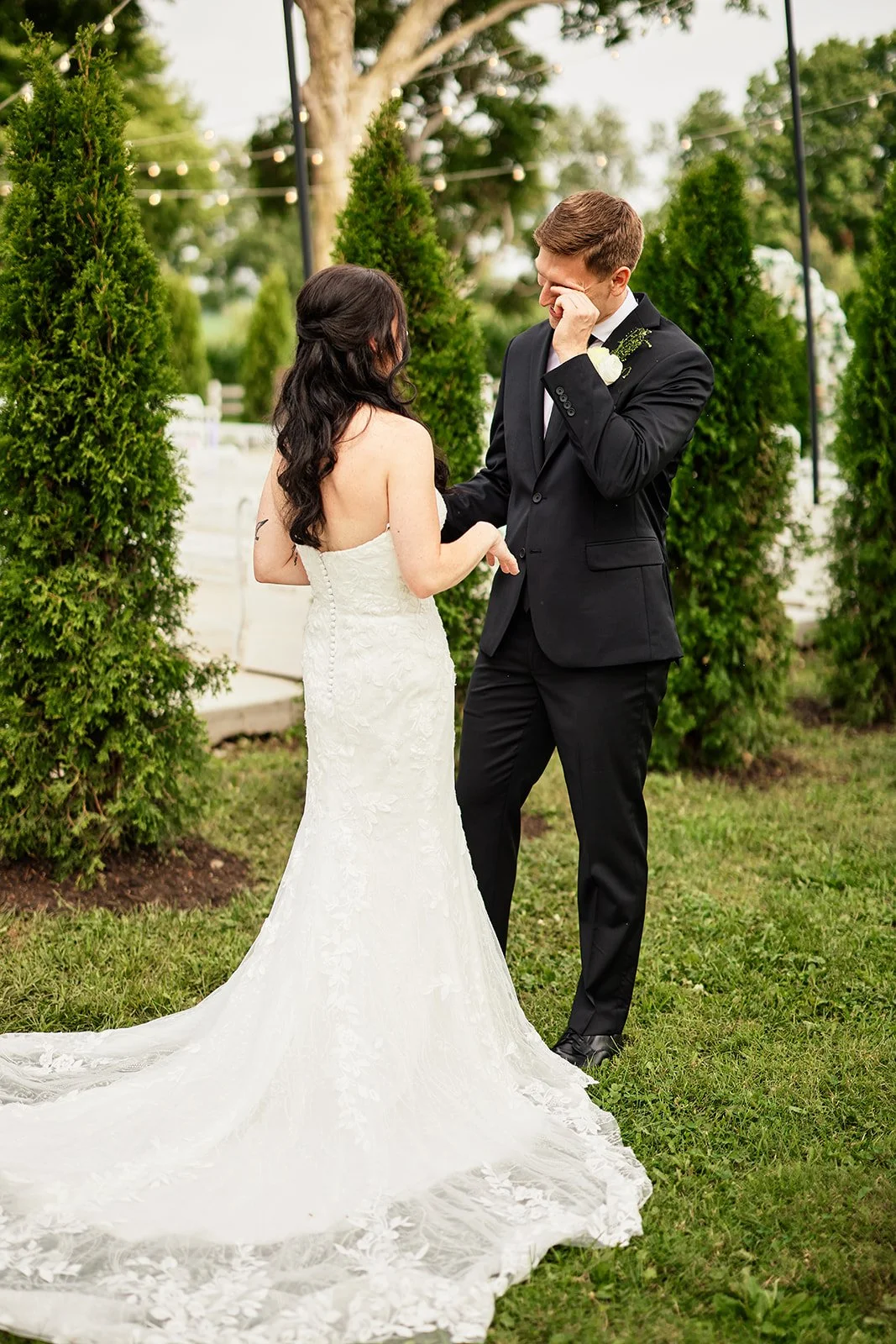 A bride and groom during their wedding ceremony outdoors. The bride is wearing a strapless white lace gown with a long train, and the groom is in a black suit with a white boutonnière. They are holding hands, and the groom is wiping tears from his ey