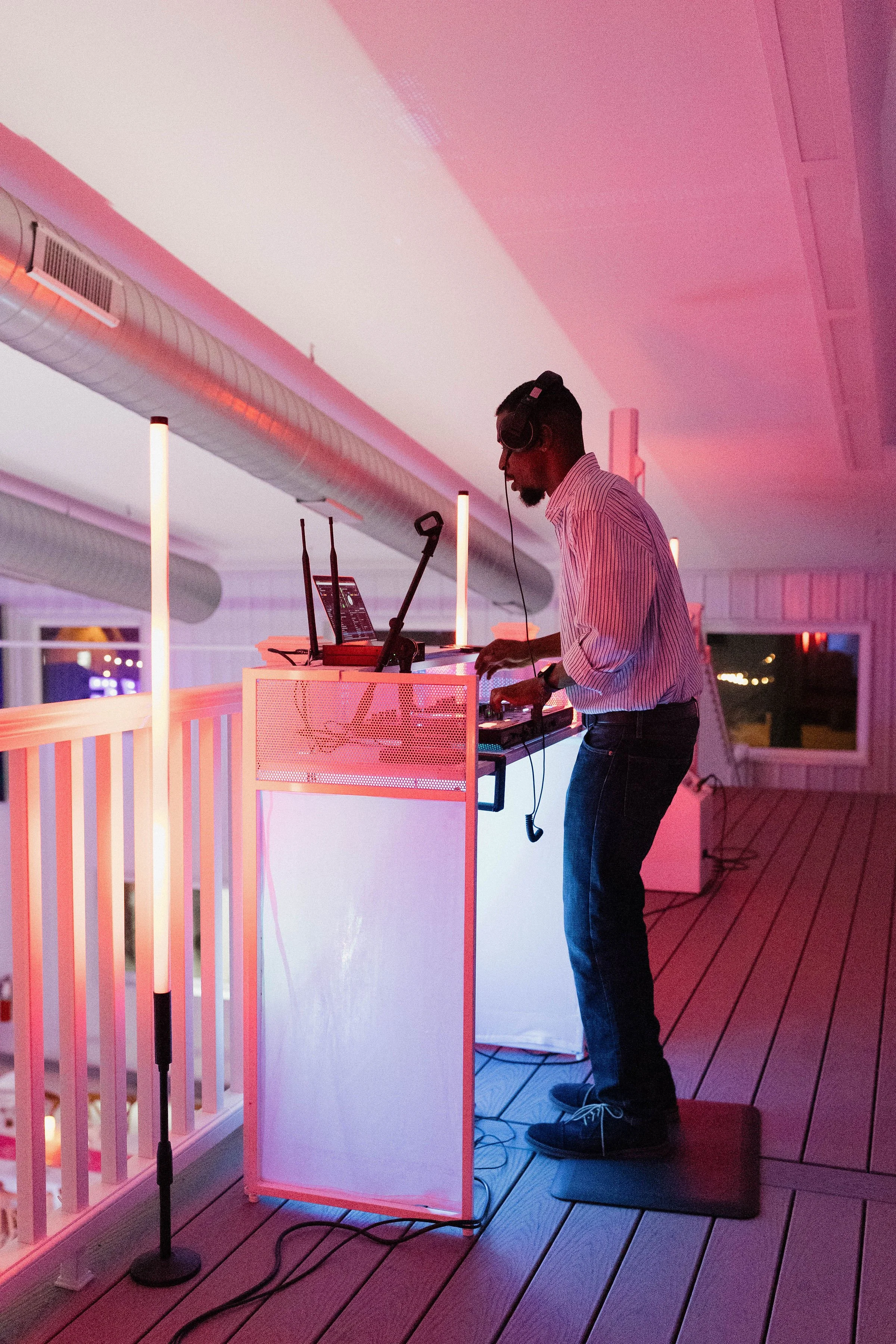 A DJ in a striped shirt and jeans wears headphones and stands on a black mat, operating DJ equipment on a white illuminated stand, with colorful pink and purple lighting in a cozy indoor venue.