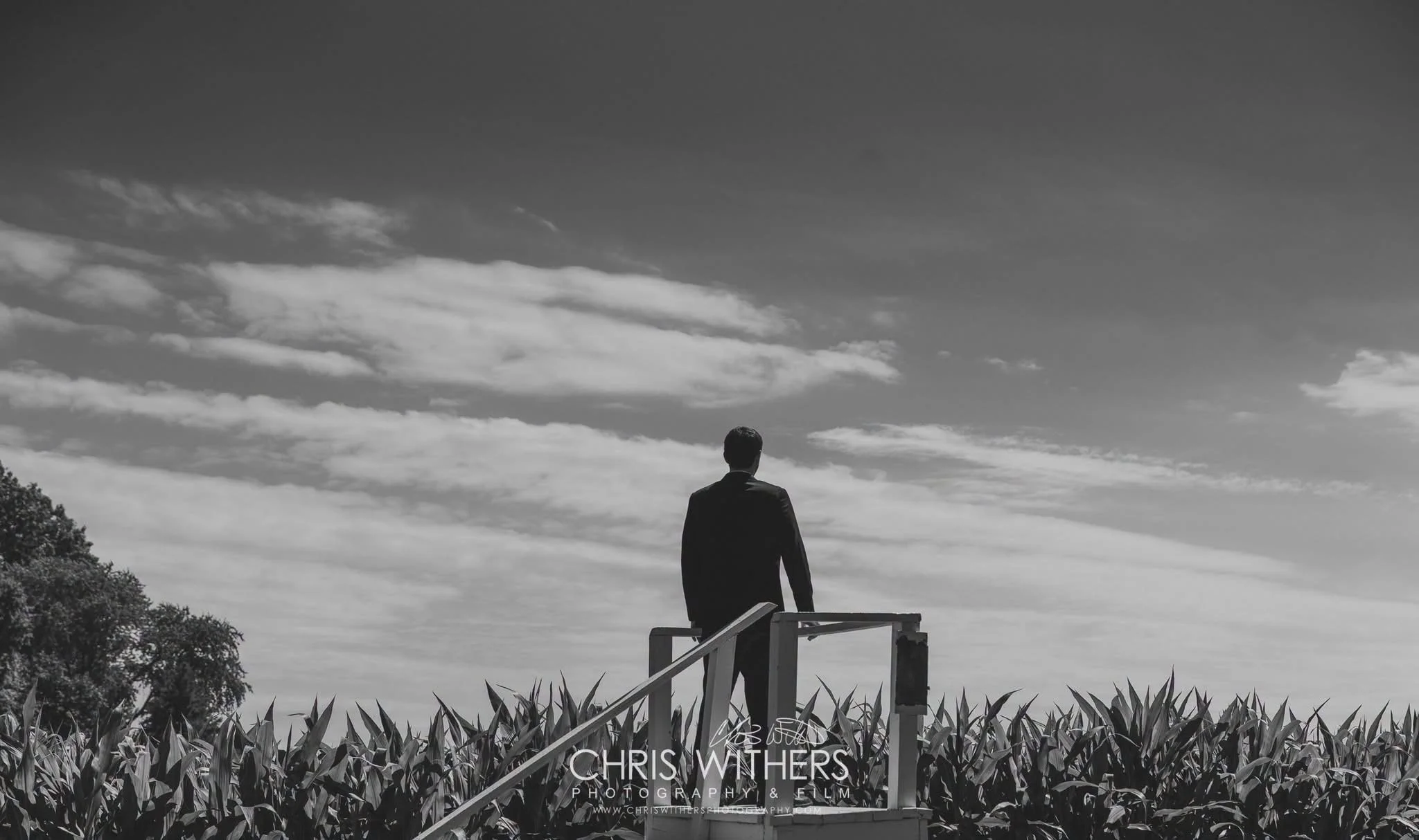 A person in a suit stands on a platform outdoors, overlooking a landscape with trees and cloudy sky, in black and white.