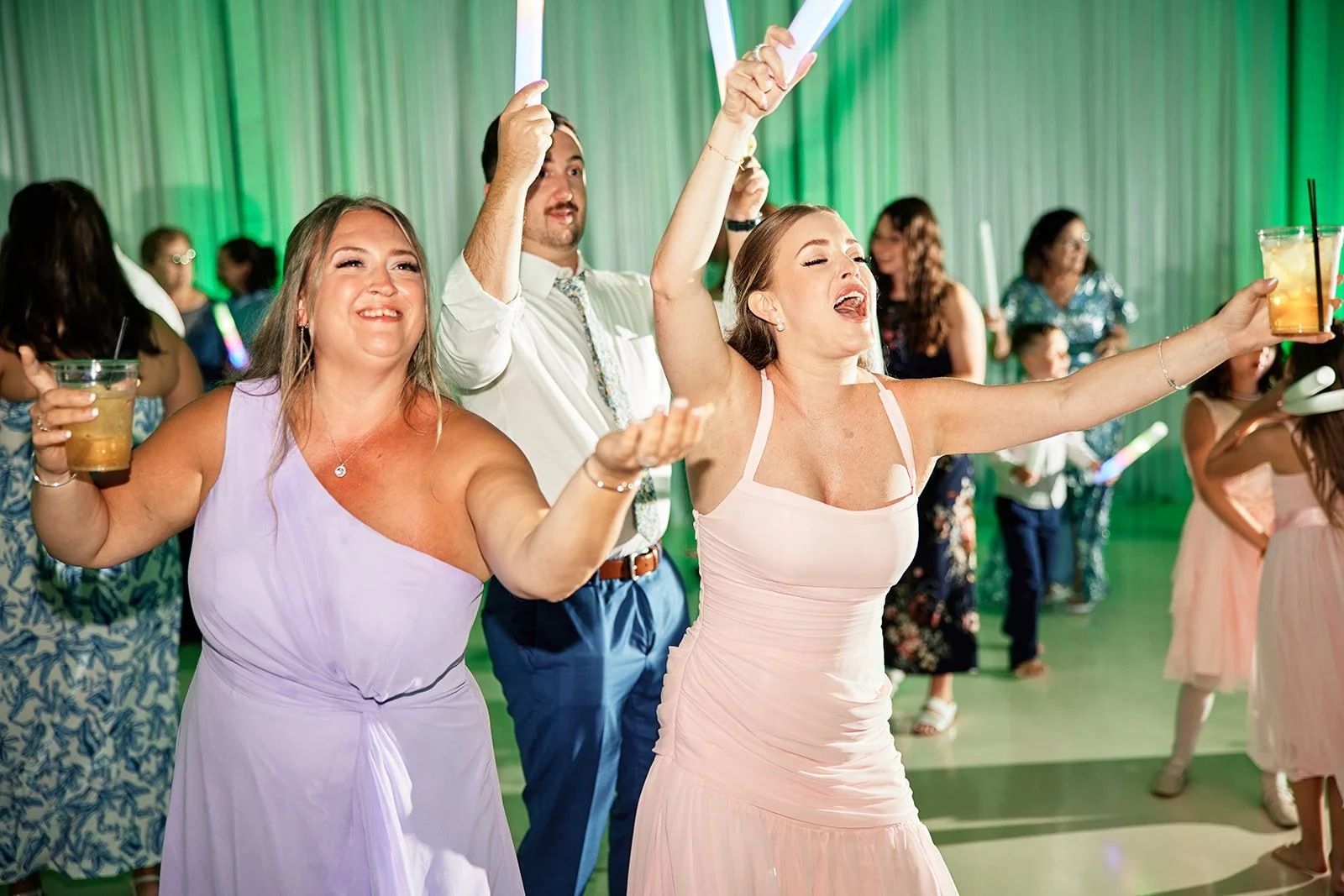 People dancing and celebrating at a party, some holding drinks, with a green-lit background. Friends, guests, bridesmaids and maid of honor.