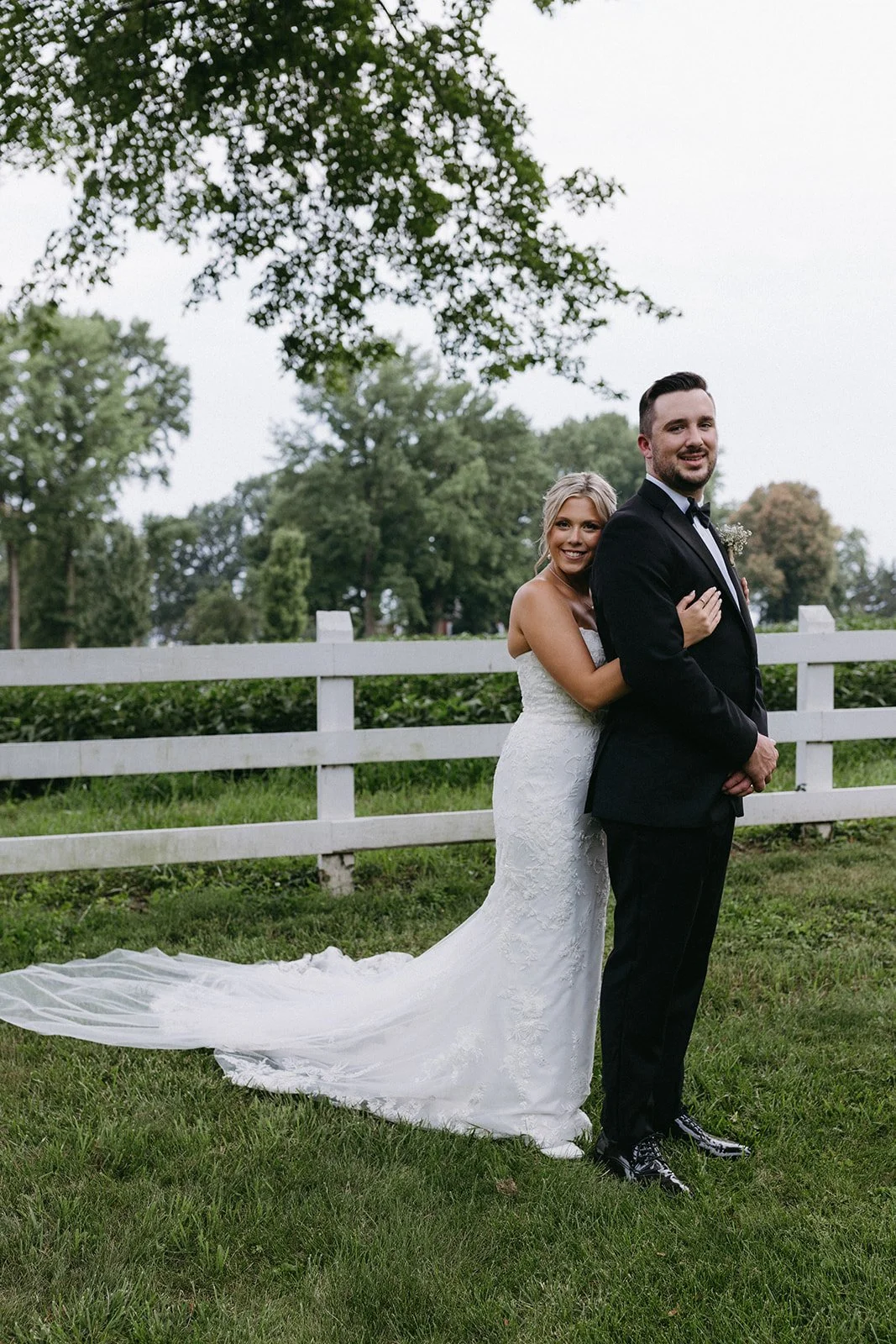 A bride and groom standing outdoors by a white fence, smiling and embracing, with trees and a cloudy sky in the background.