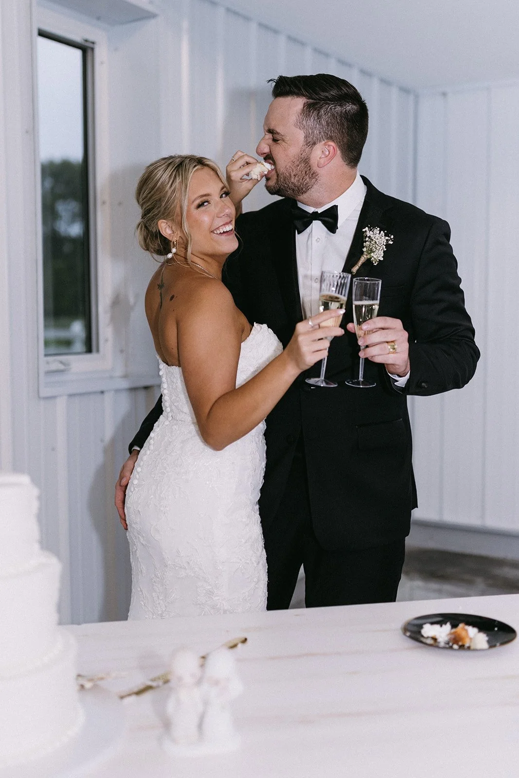 A newlywed couple in wedding attire sharing a joyful moment, holding champagne glasses with a cake and dessert plate in the foreground, inside a white-paneled room.