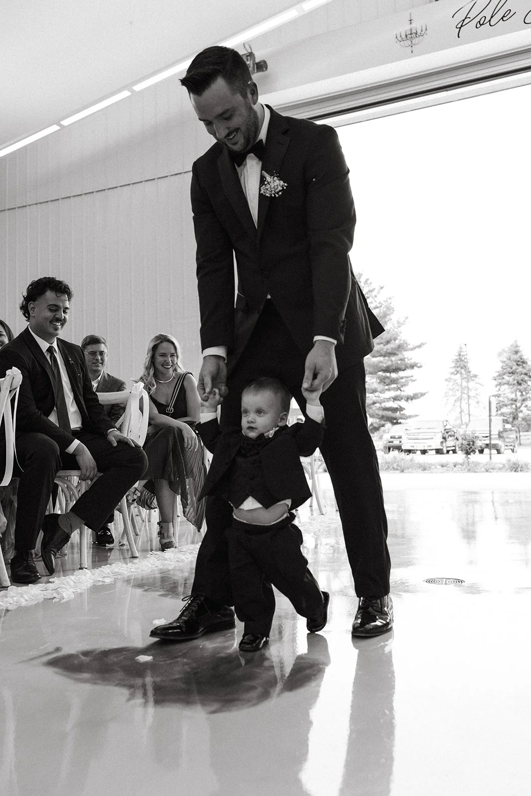 A man in a tuxedo holding a young boy by the hands, walking on a reflective floor in a decorated venue (the PBC Venue) with seated guests smiling and watching.