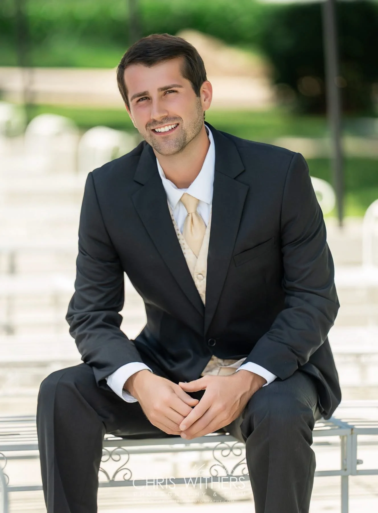 A young man in a black suit and light-colored tie sitting on a park bench outdoors, smiling at the camera, with green trees and a pathway in the background.