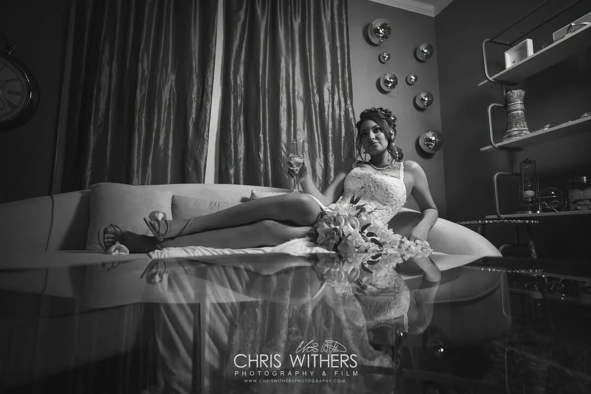 A woman in a wedding dress reclining on a sofa, holding a glass of champagne, with a bouquet of flowers on the sofa.