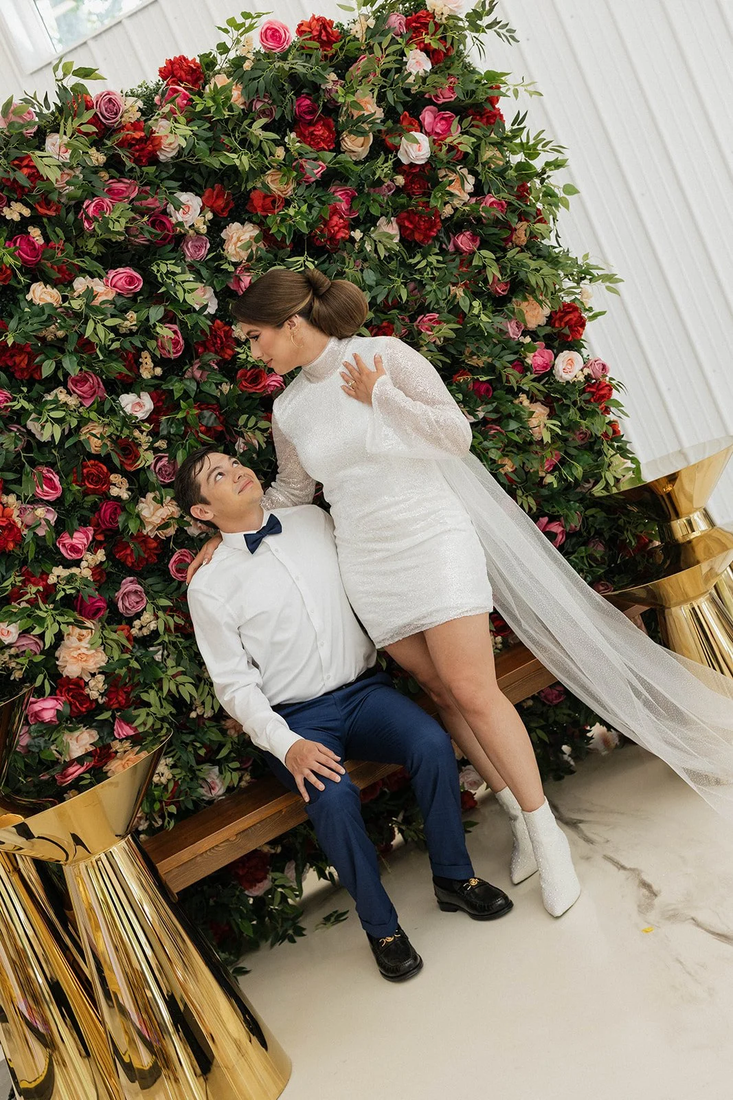 A man in a suit and woman in a reception dress with veil, sit on a brown bench in front of a rose wall and gold cake pillers.