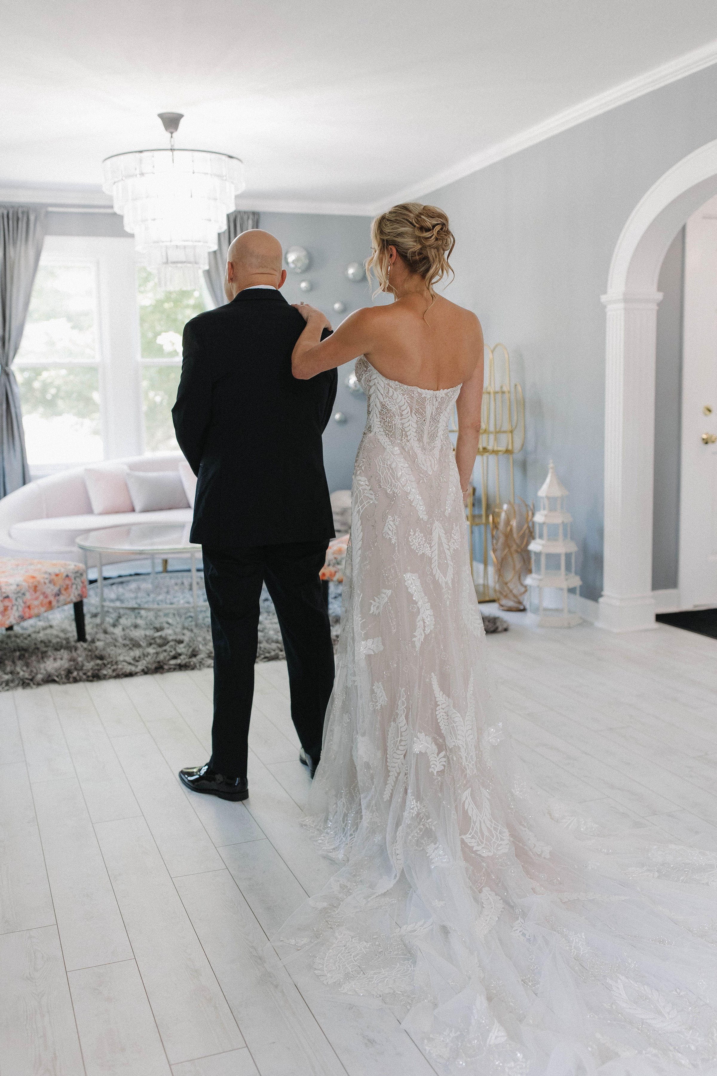 A bride in a white wedding dress standing next to a man in a black suit in a bright living room, with sunlight coming through large windows, during a wedding moment.