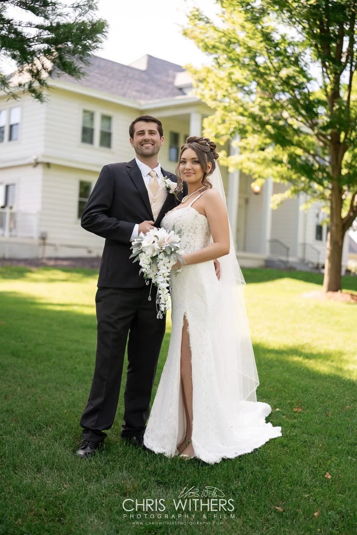 A bride and groom standing on a grassy lawn in front of a light-colored house with trees around them. The groom is wearing a black suit, and the bride is in a white wedding dress holding a bouquet of flowers.