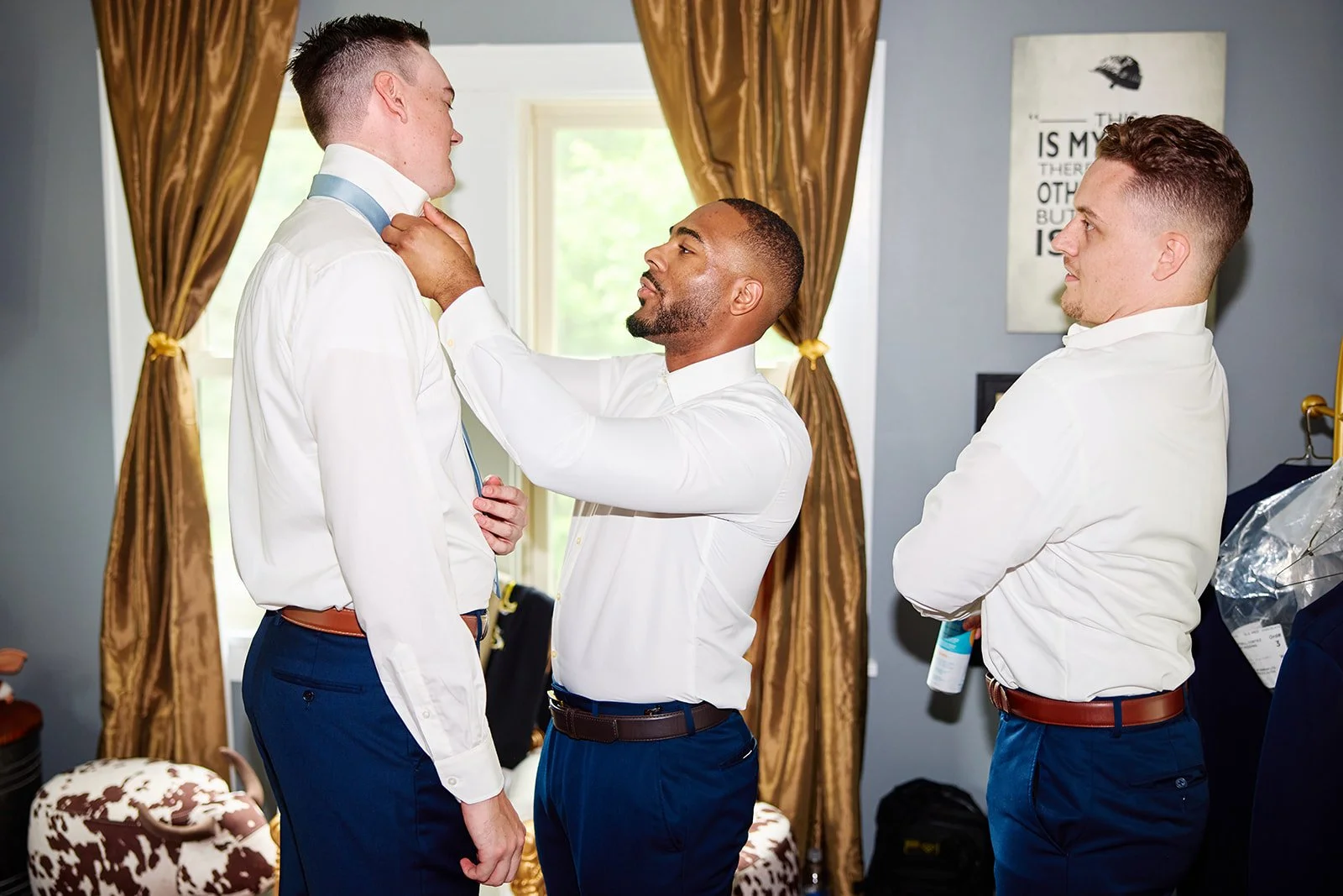 Three men in white shirts and blue pants preparing for a formal event. One man adjusts the tie of another, while the third man watches, holding a bottle. The room has gold curtains and a framed picture on the wall.