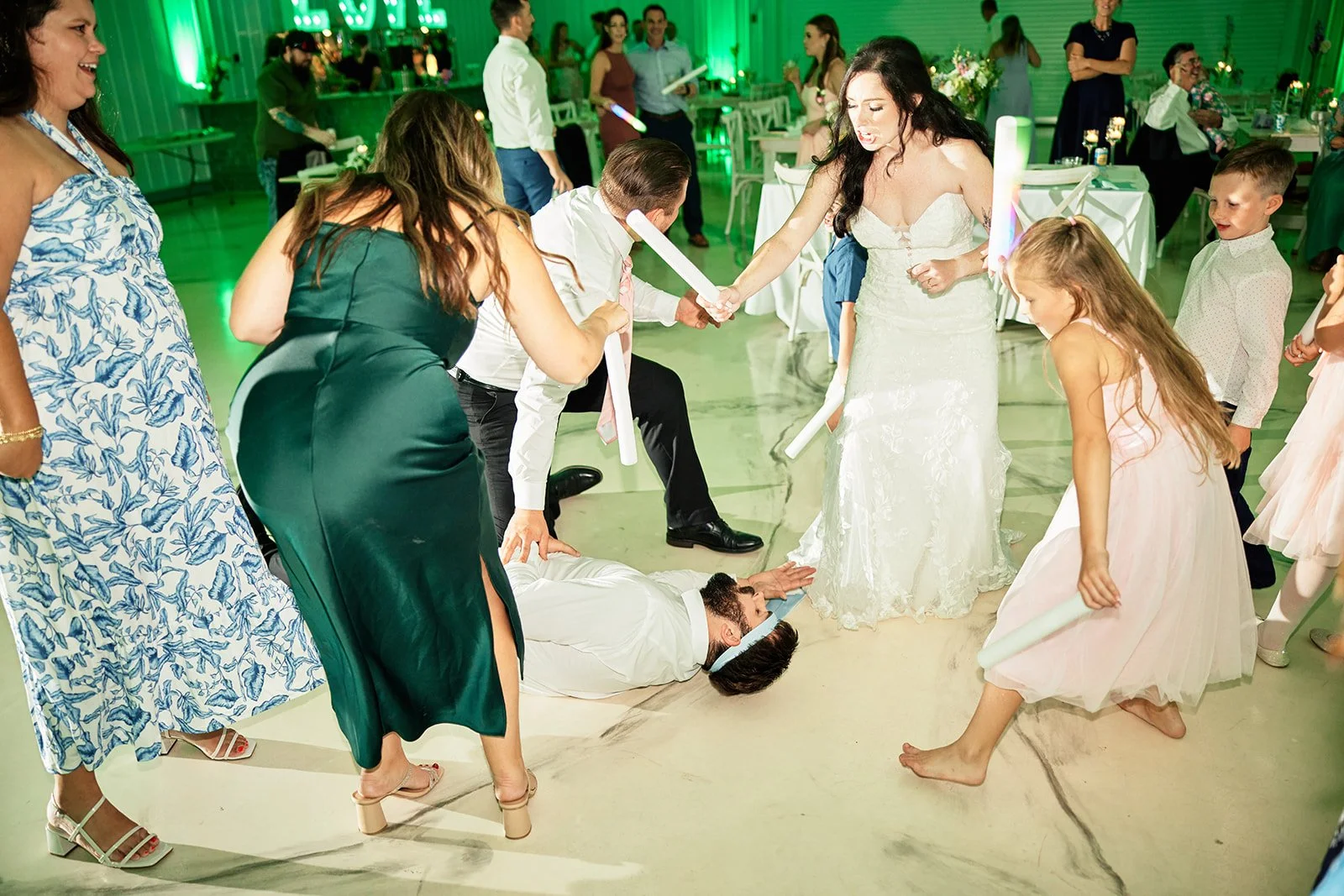 Guests at a wedding reception playing a game where a man is lying on the floor with a woman standing over him, and others surrounding them holding glow sticks, with a green-lit decorated background.