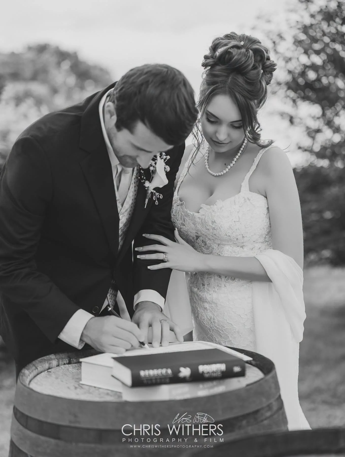 A black and white photo of a bride and groom during their wedding ceremony. The groom is signing a document on a table, while the bride looks on with her hand resting on his forearm. The bride is wearing a lace wedding dress and pearl necklace, and h