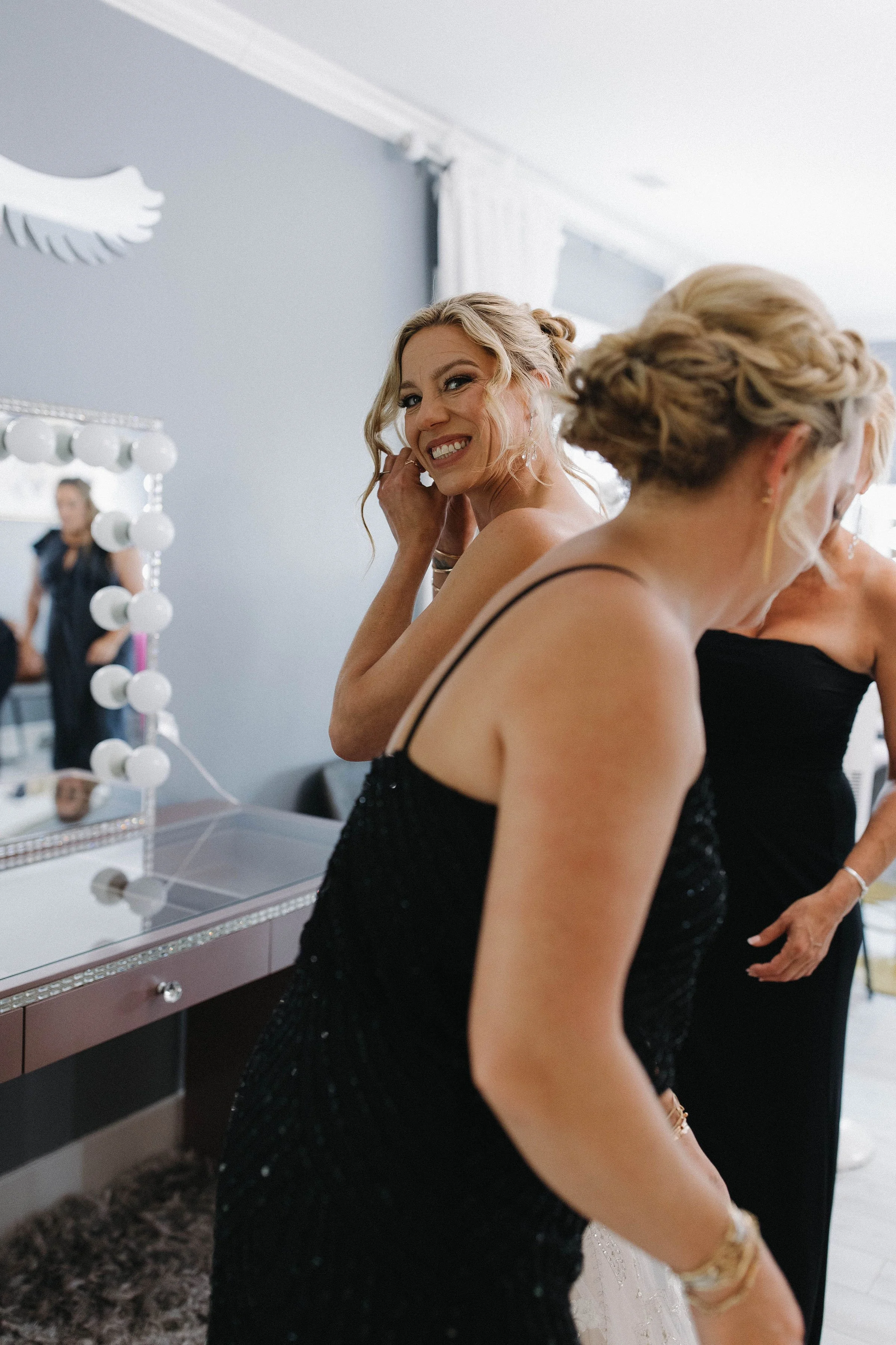 Two women getting ready, with one adjusting her hair and both dressed in elegant black dresses, in a room with a mirror and vanity lights.
