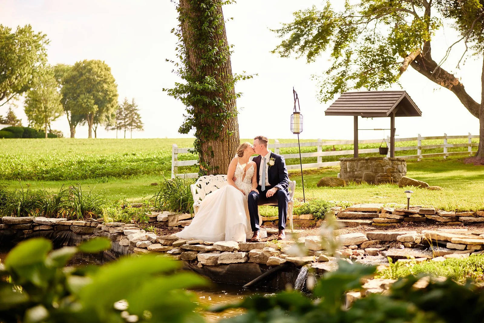 A bride and groom sit on a white bench kissing in a scenic outdoor garden with green grass, trees, and a small pond or stream in the foreground.
