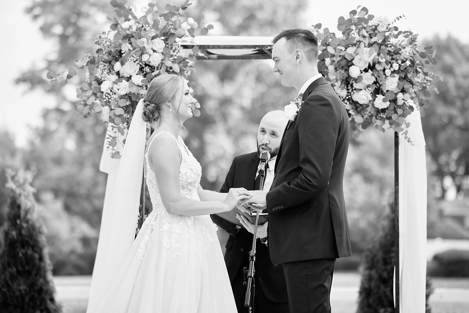 A black-and-white photo of a wedding ceremony with a bride and groom holding hands, facing each other under an arch decorated with flowers, with an officiant standing behind them.