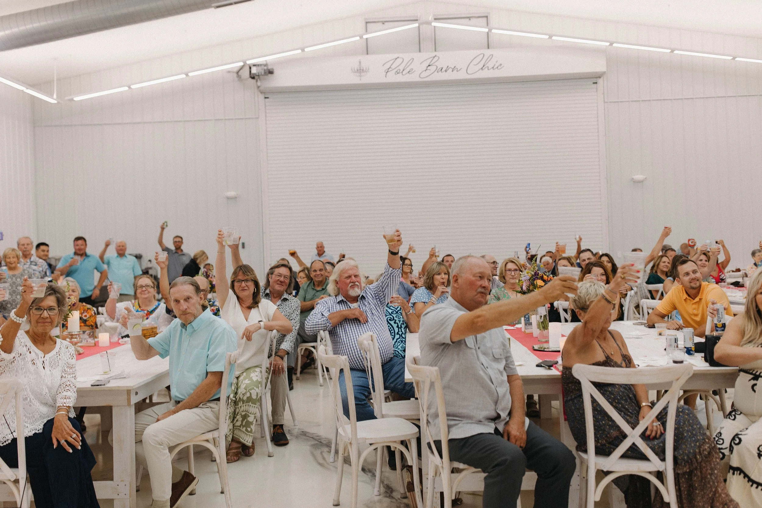 People sitting at tables inside a large, white event space with a sign that reads 'Pole Barn Chic.' Guests are raising glasses in a toast, with some smiling and others looking forward.