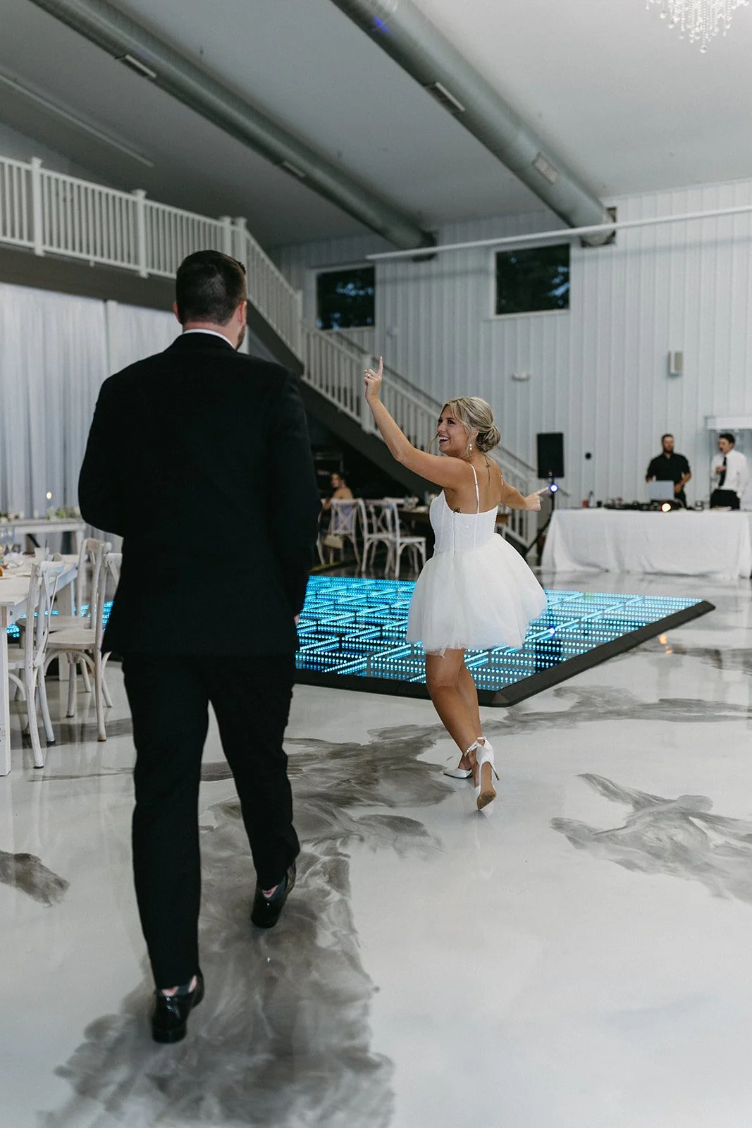 A bride in a white dress dancing joyfully on a lit-up dance floor at a wedding reception, with a groom in a black suit watching her.