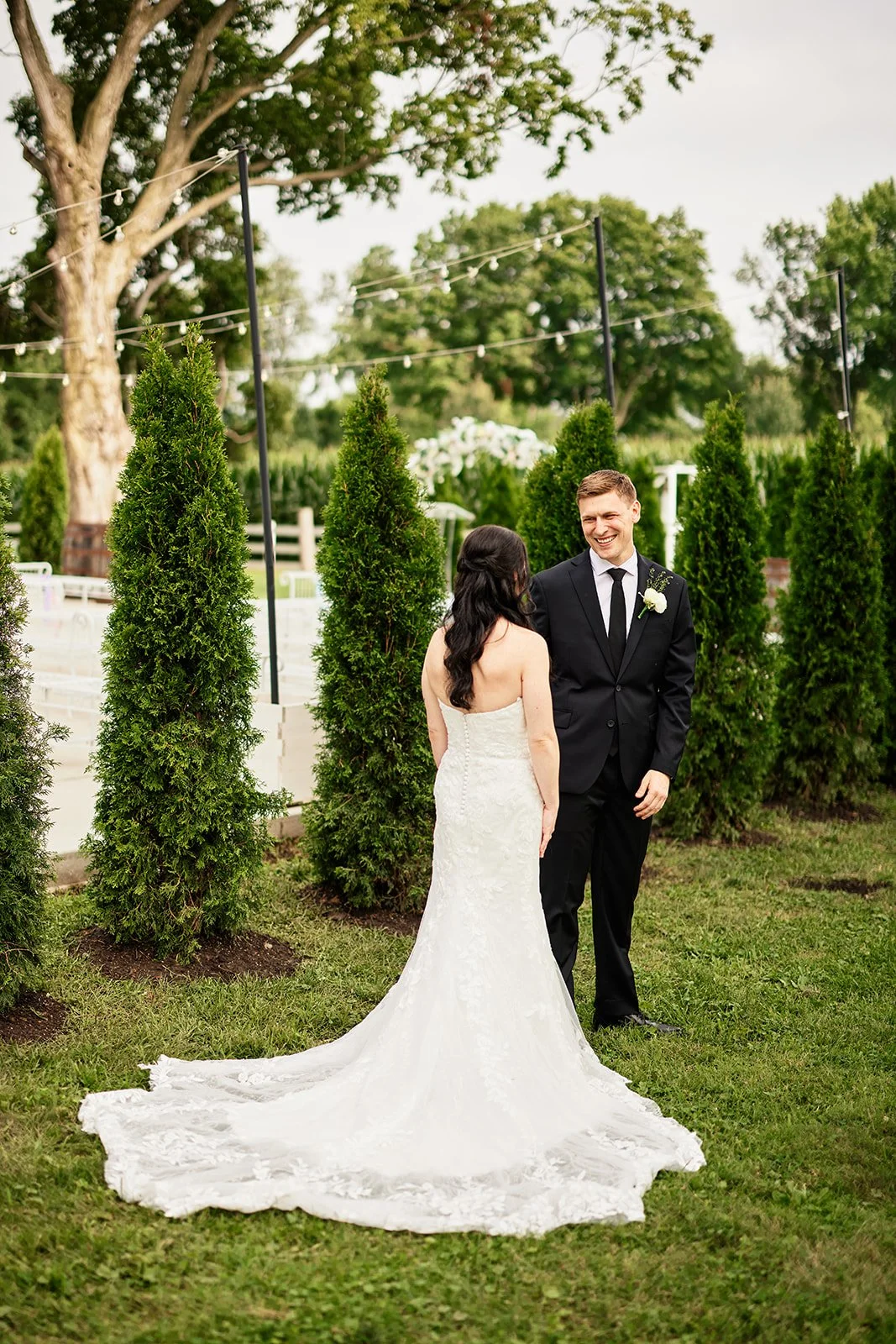 A bride and groom standing outdoors during a wedding ceremony. The bride is in a white strapless wedding gown with a train, and the groom is in a black suit with a white shirt and black tie. They are smiling at each other, surrounded by green trees a