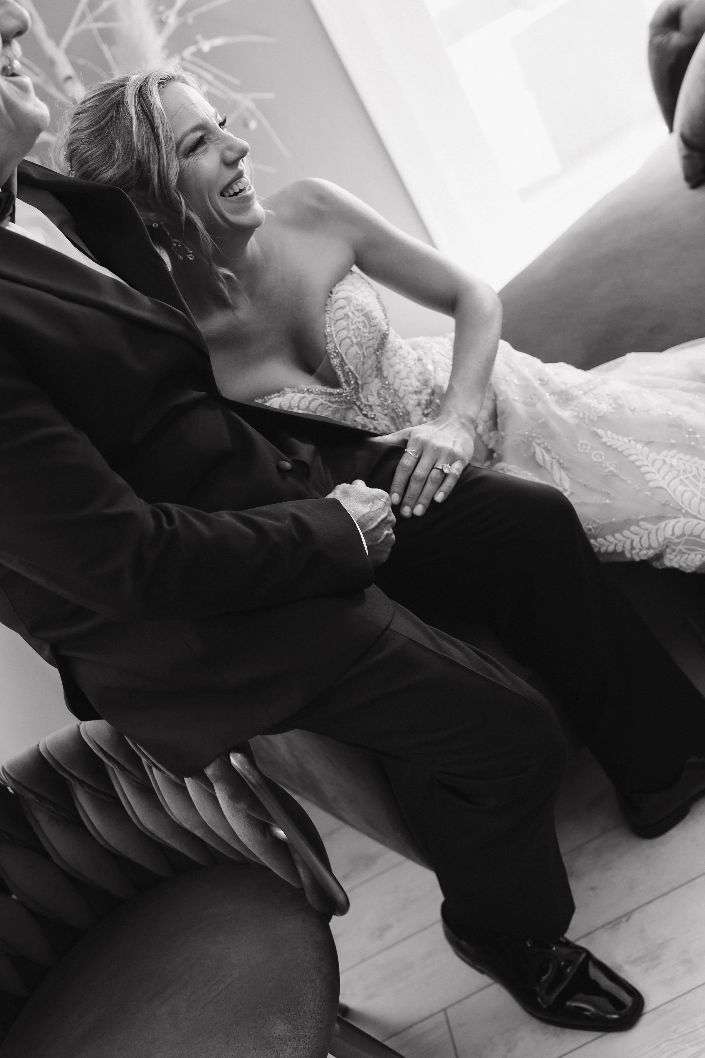 A woman in a wedding dress laughing while sitting beside a man in a suit holding hands.