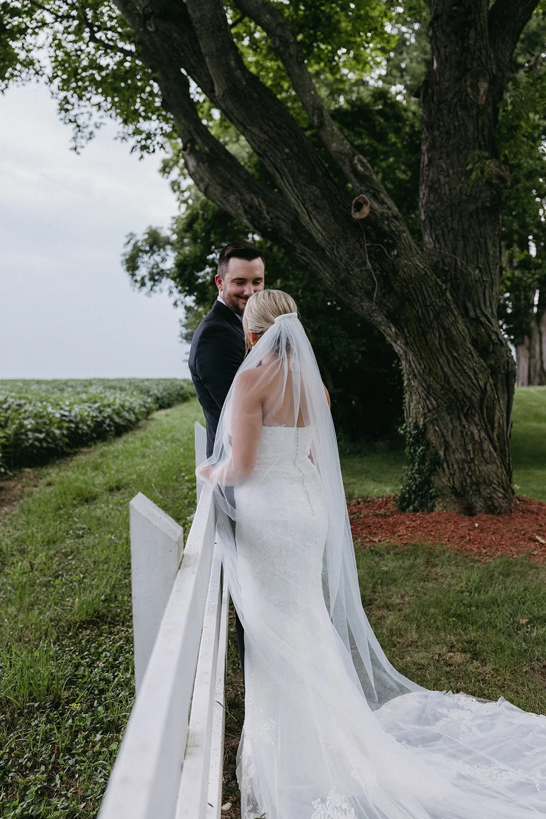 A bride in a white wedding dress with a long veil and a groom in a dark suit standing close together outdoors near a large tree, smiling at each other on a grassy area.