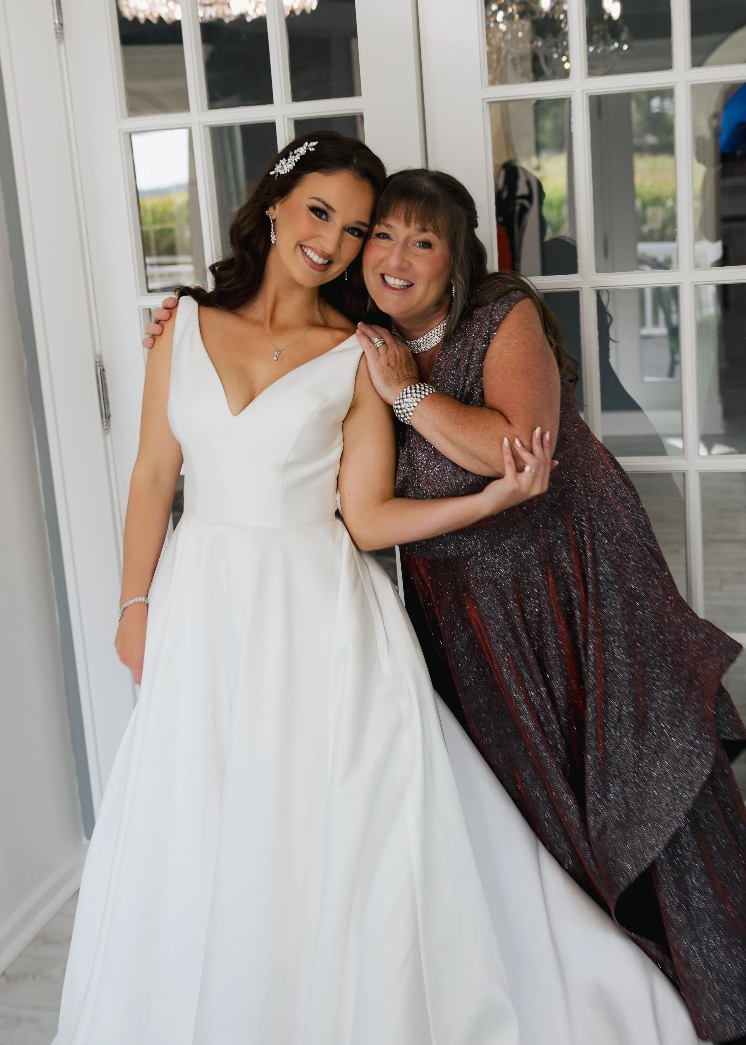 A smiling bride in a white wedding dress and her mother in a sparkly dress pose together inside near glass doors. The bride has dark hair with a decorative hairpiece and jewelry. The mother has shoulder-length hair and jewelry, and they are both smil