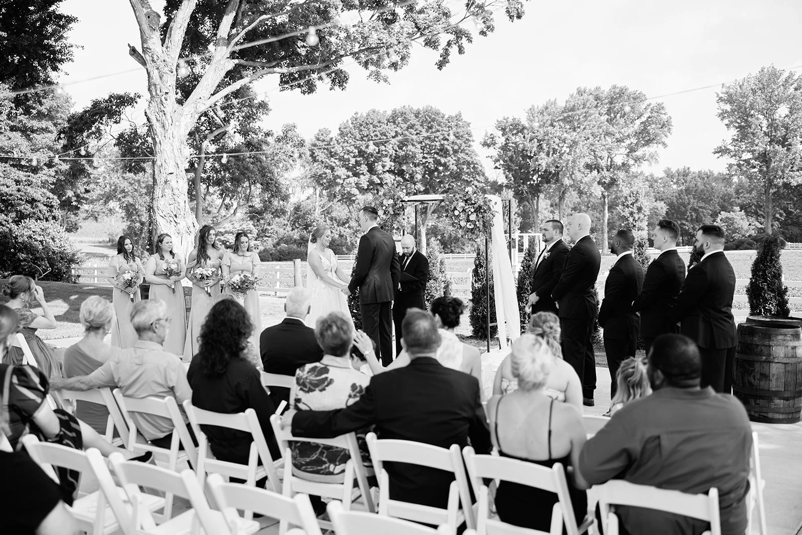 A black and white outdoor wedding ceremony with the bride and groom holding hands at the altar, surrounded by guests seated in chairs, and bridesmaids and groomsmen standing on either side under large trees.