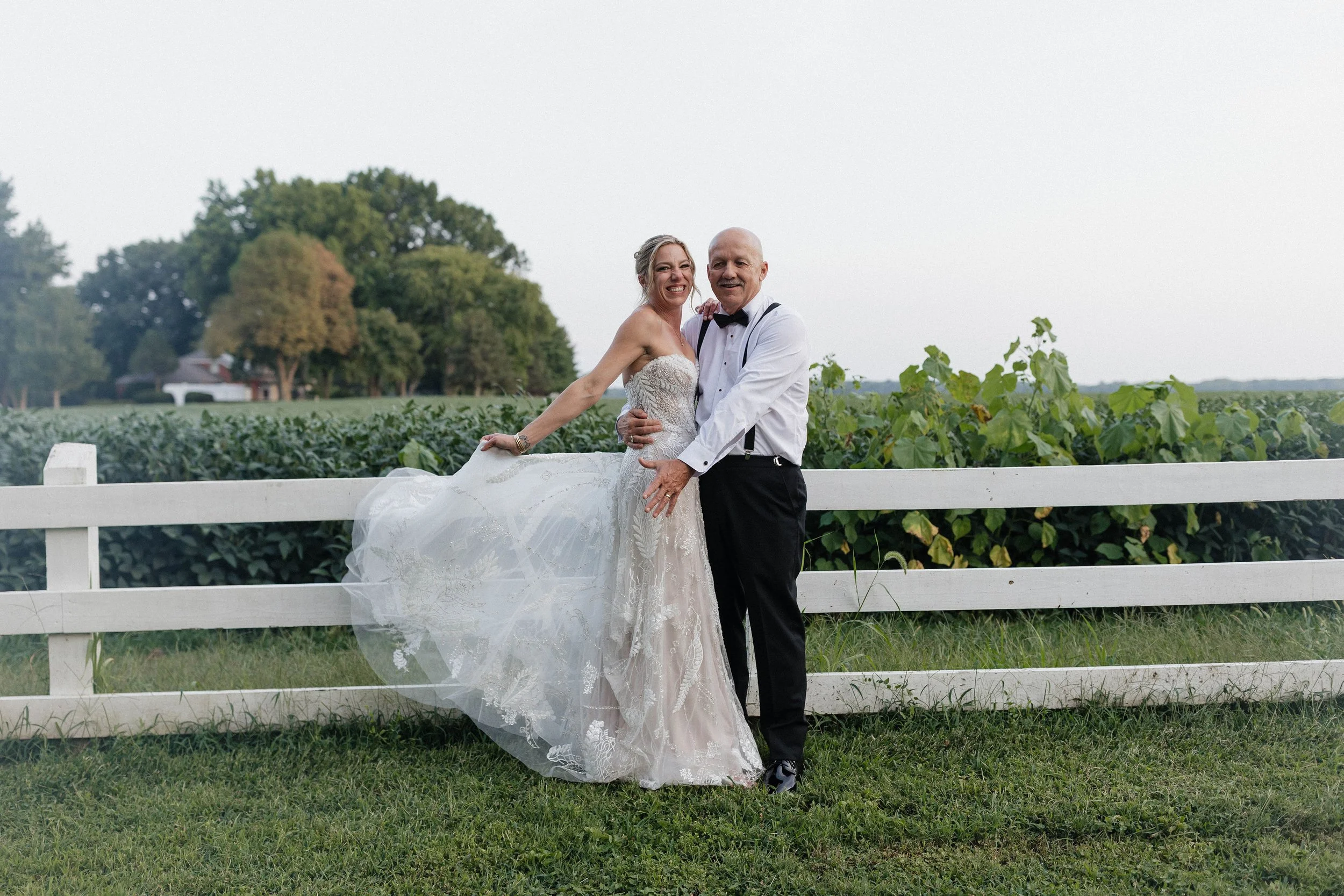 A woman in a wedding dress and gentleman in formal attire are smiling and posing in front of a white fence with a green field and trees in the background.
