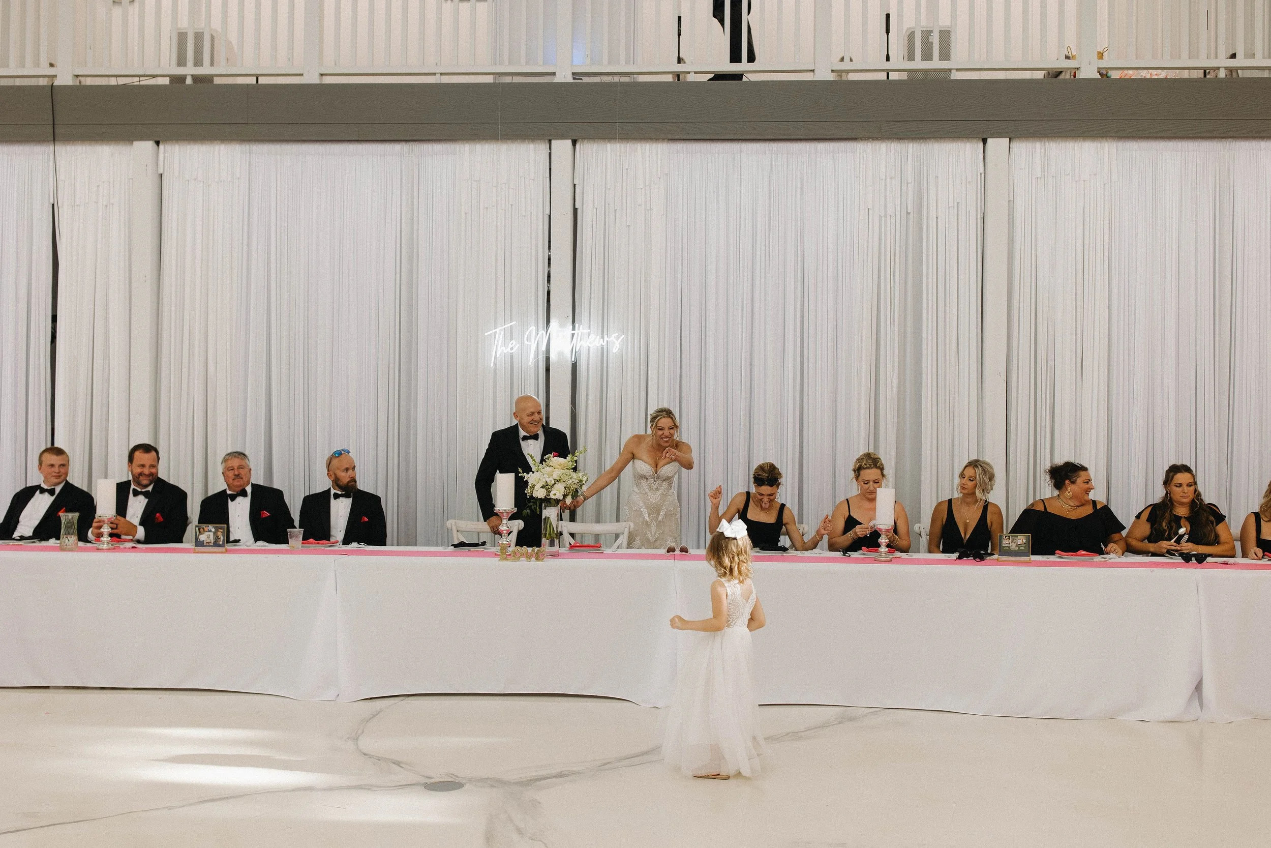 A bride and groom at their wedding reception are holding hands across a long table with bridesmaids and groomsmen. The bride is smiling while the groom is smiling and looking at a young girl in a white dress with a bow in her hair, who is standing in