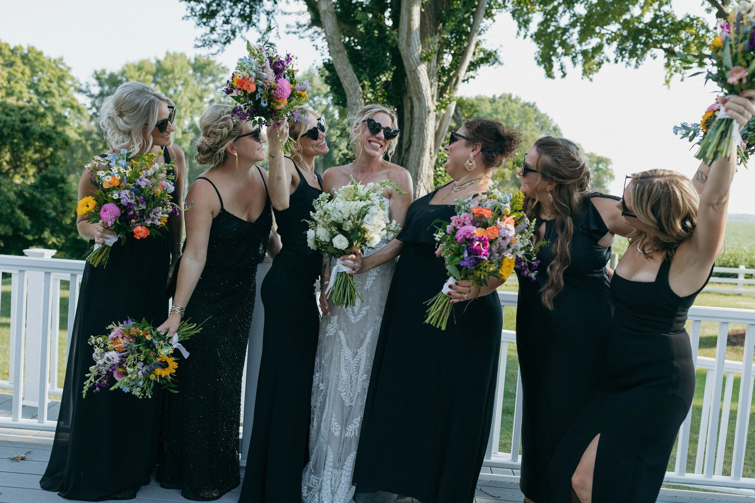 A group of women in black dresses and sunglasses celebrating outdoors at a wedding, with a bride holding a bouquet of white flowers, surrounded by bridesmaids holding colorful bouquets, standing on a deck with a white railing under a tree.