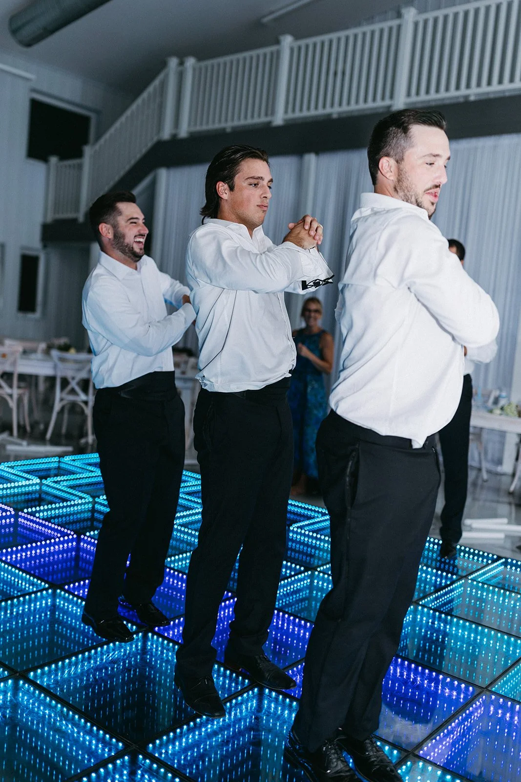 Three men in white shirts and black pants dancing on a grid of LED-lit mirrors at a party, with a woman in a blue dress watching in the background.