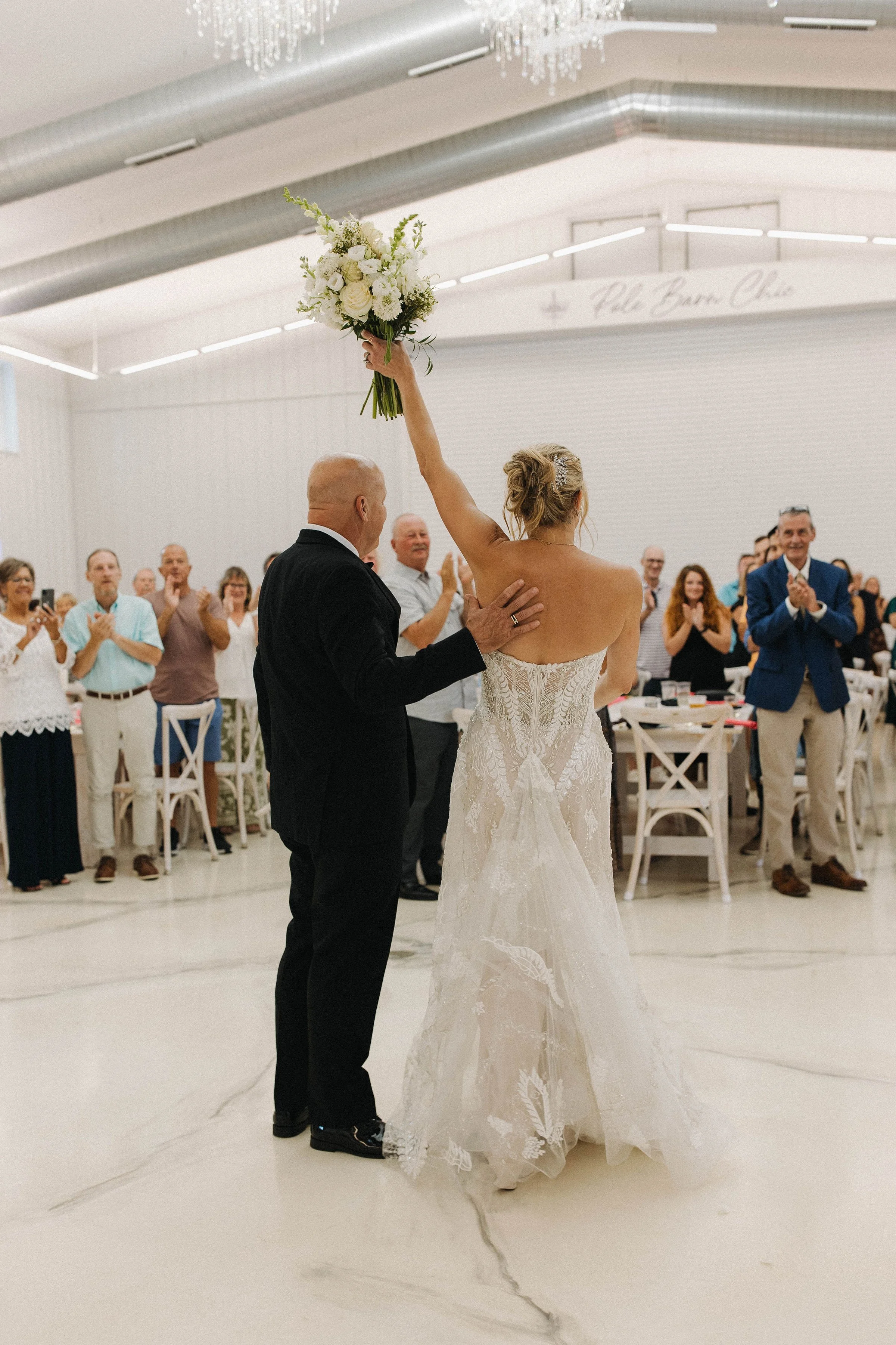 Bride holding a bouquet above her head as she walks down the aisle with her father, surrounded by smiling and clapping guests at a wedding reception.