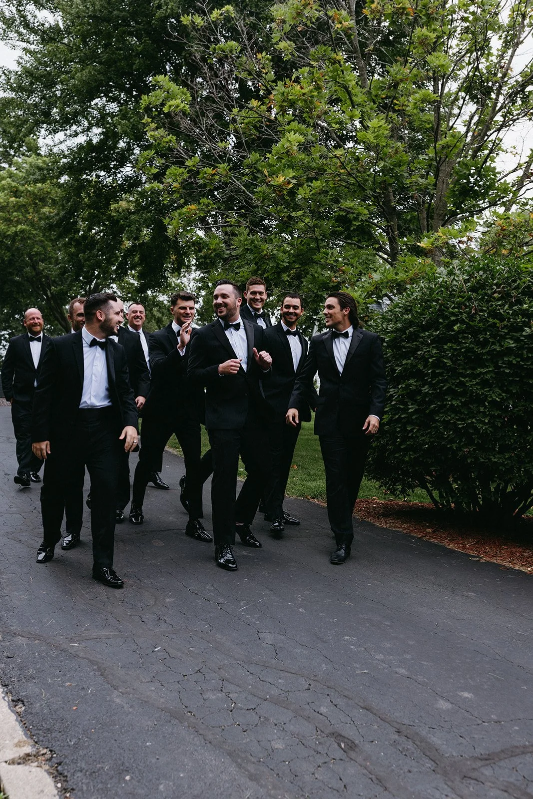 A group of men in tuxedos walking and smiling on a paved path outdoors, surrounded by green trees and bushes.