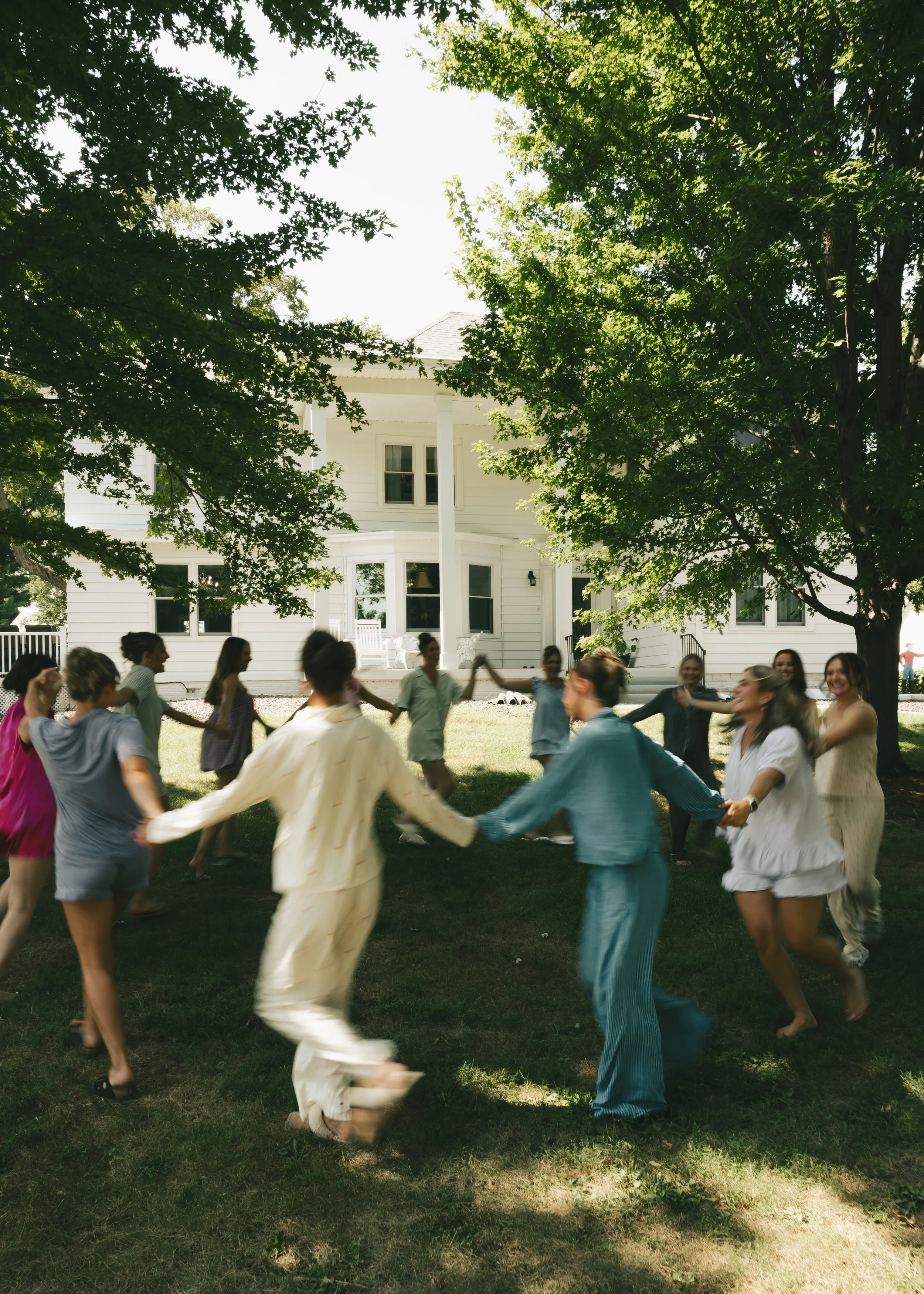 Group of people holding hands in a circle in a backyard with trees and a white house in the background.