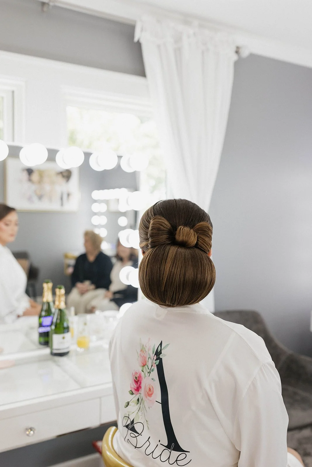 Bridal prep room with a woman wearing a white robe that has a floral and 'bride' logo on the back, styled hair, and other women in the background reflected in a mirror.