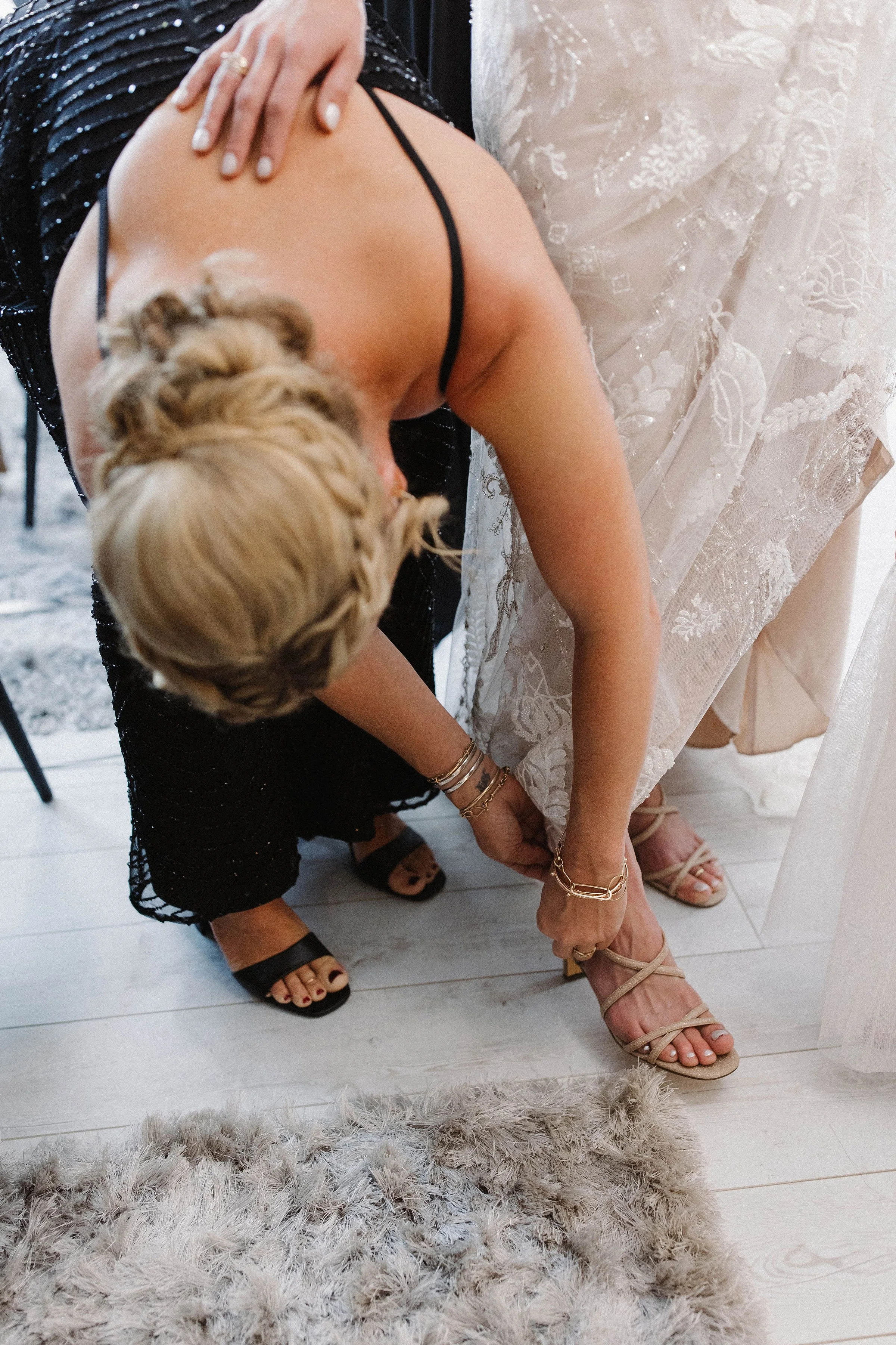 A woman in a black dress helps another woman in a white wedding gown put on beige high-heeled sandals, bending over to assist.