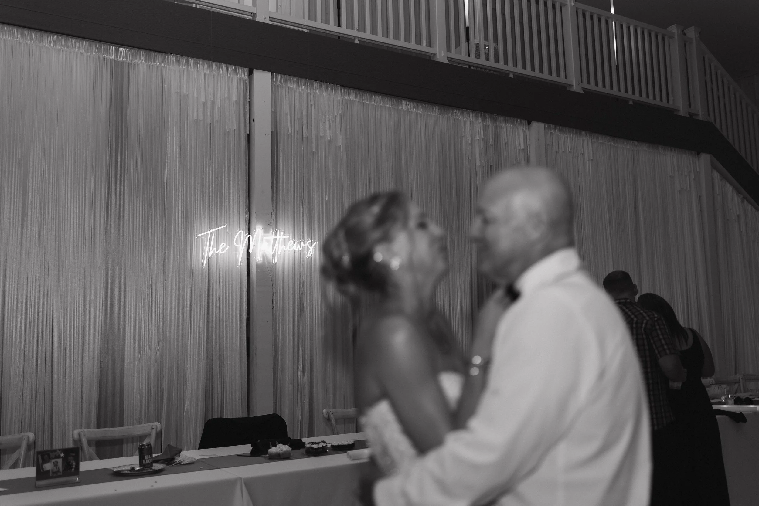 A wedding reception with an older couple dancing, blurred in the foreground, against a wooden wall with a neon sign that reads 'The Matthews'.