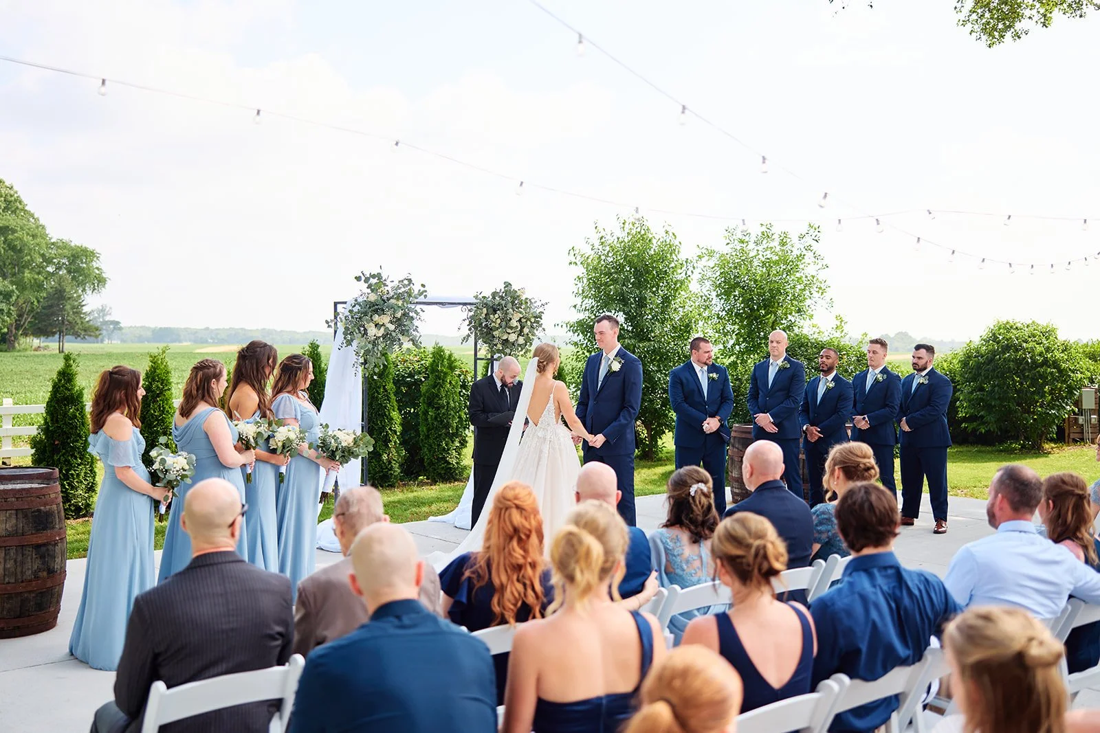A wedding ceremony outdoors with the bride, wearing a white gown, and groom, in a navy suit, holding hands and facing each other under a floral arch. Bridesmaids in light blue dresses and groomsmen in navy suits stand beside them. Guests sit on white